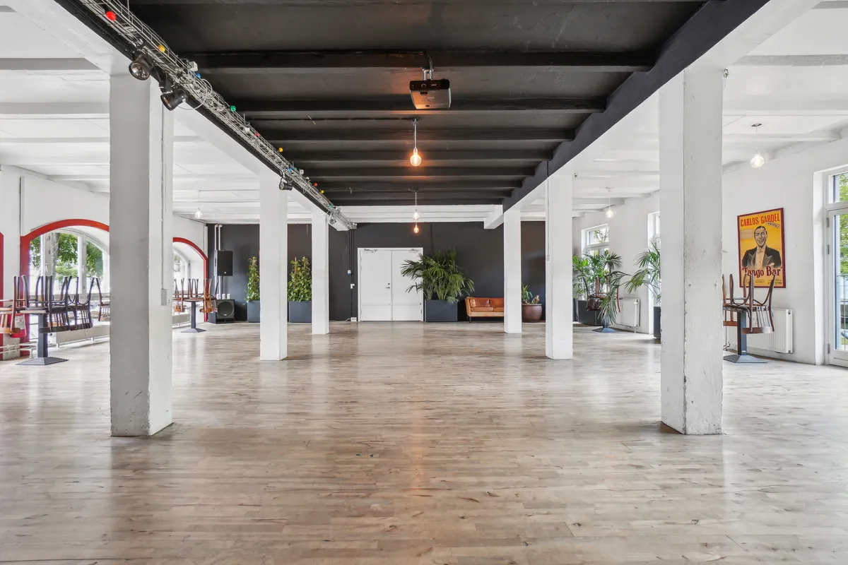 Store værelse med højt loft, lyse loftbjælker, hvidt gulv og lysekroner. Der er store ruder, planter og en projektor i loftet. Der er også en skilt med en mand på.