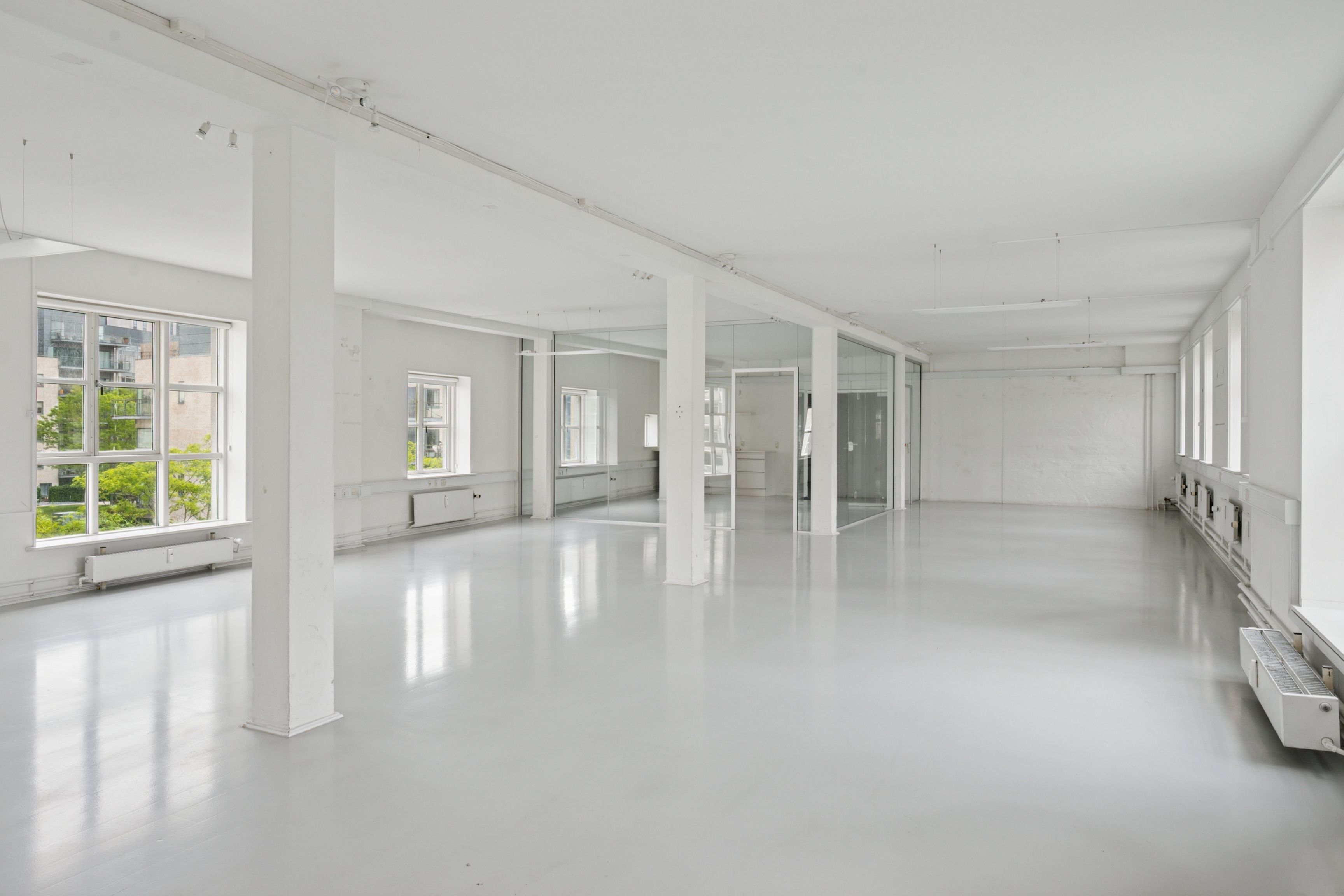 Forslag til alt-tekst: En stor og rummelig kontorlokal i industriel stil med høje lofter, lyse gulve og store ruder, som reflekterer lyse fra vinduet.