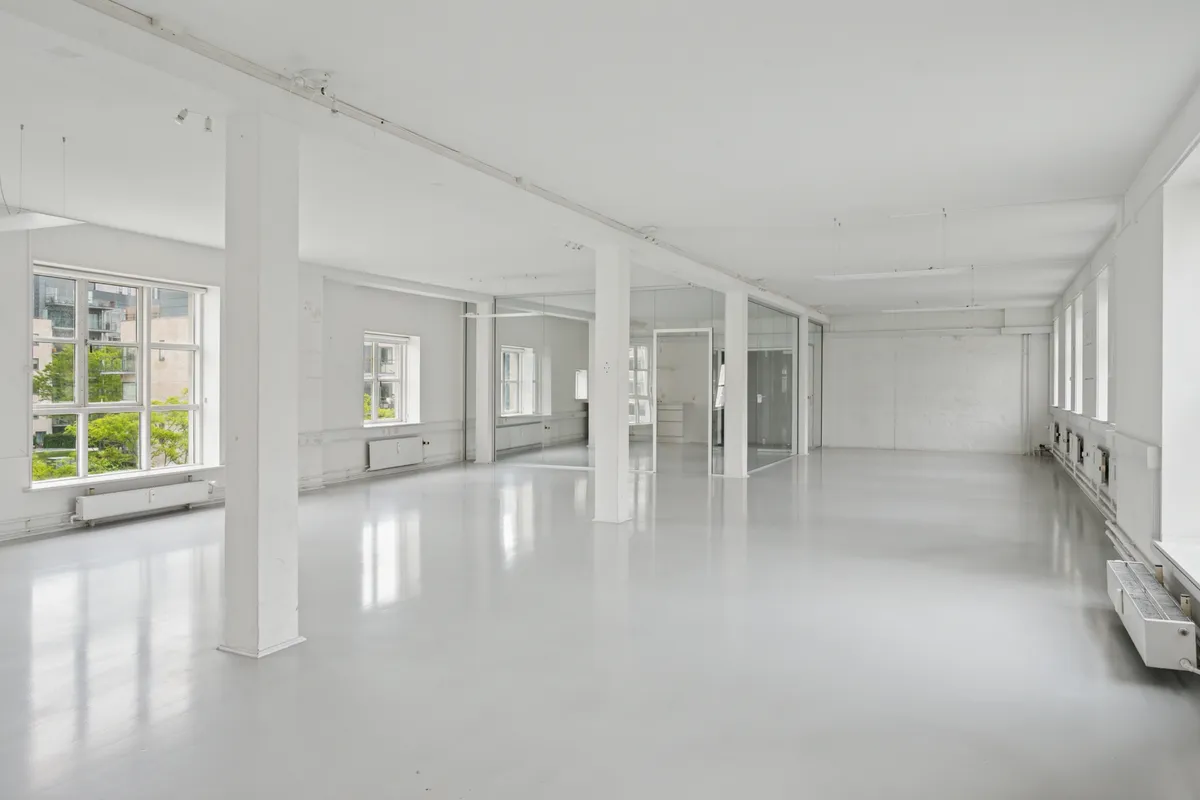 Forslag til alt-tekst: En stor og rummelig kontorlokal i industriel stil med høje lofter, lyse gulve og store ruder, som reflekterer lyse fra vinduet.
