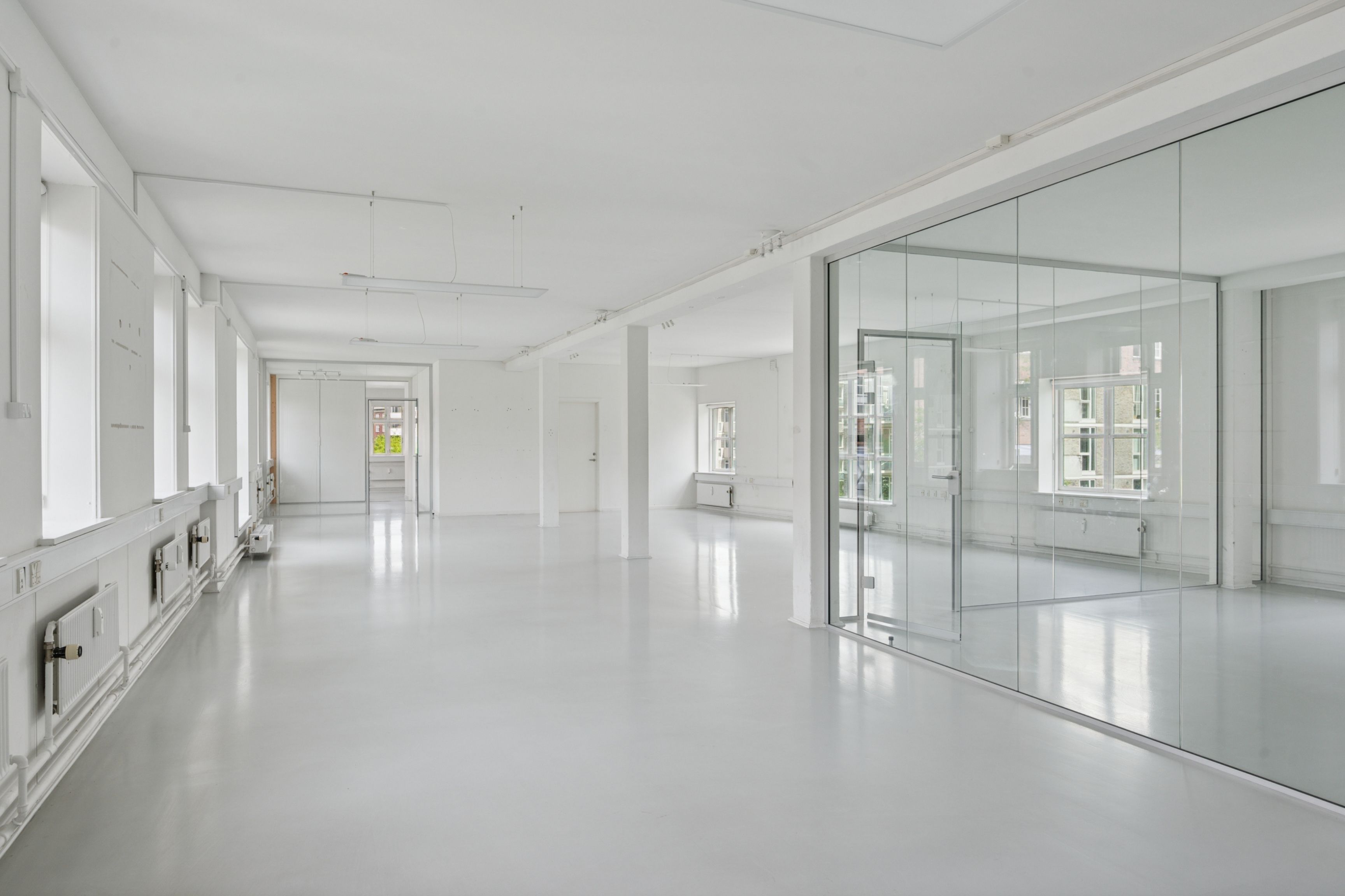 Et hulrum med hvide vægge, glaspartier og moderne gulvbelægning. Store vinduer og åbne døre giver meget lys. Et tæt erhvervsområde med flere bygninger i baggrunden.
