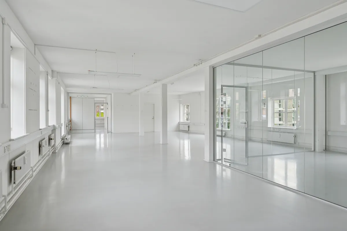 Et hulrum med hvide vægge, glaspartier og moderne gulvbelægning. Store vinduer og åbne døre giver meget lys. Et tæt erhvervsområde med flere bygninger i baggrunden.