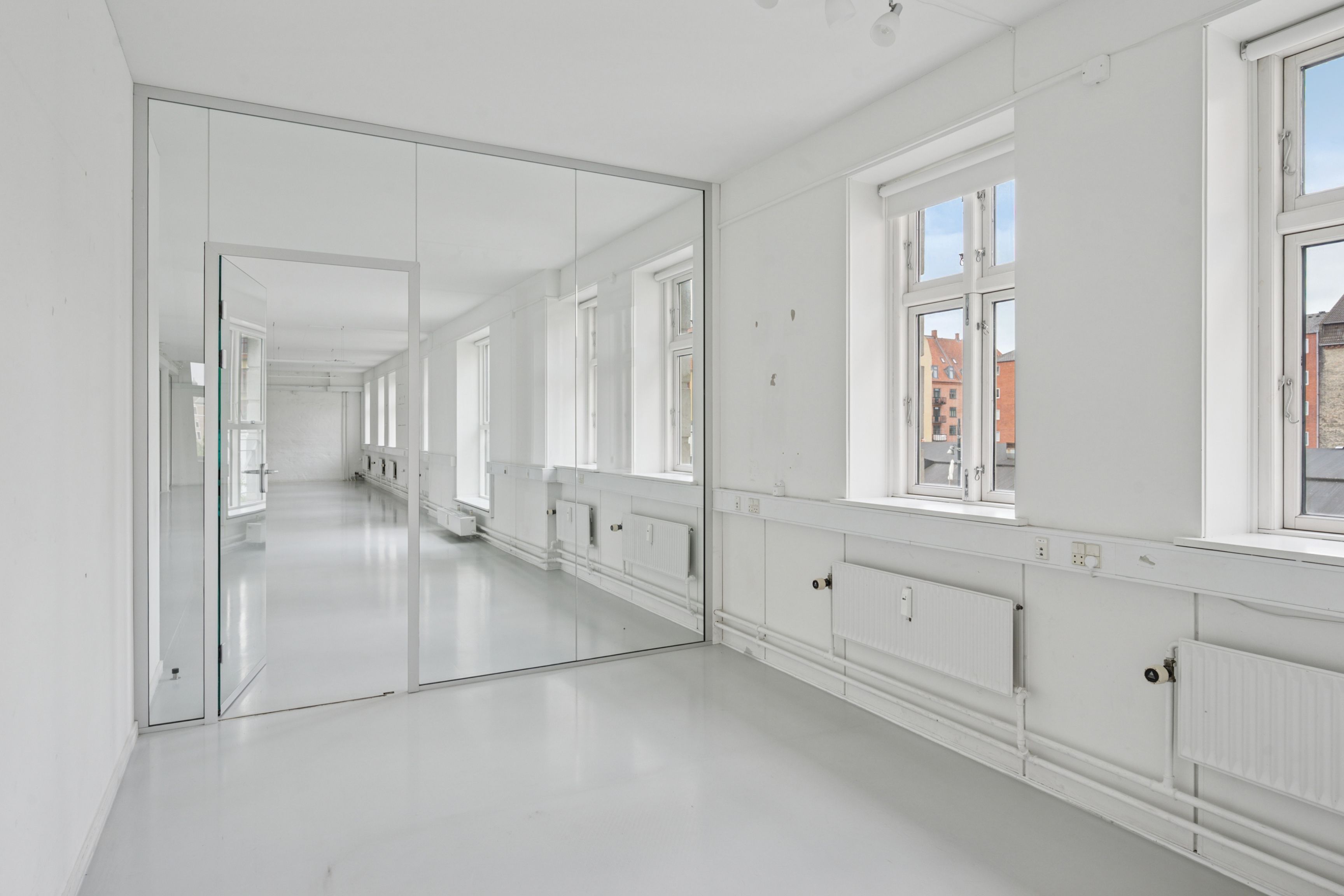 En lys og åben kontorry, med spejl og hvide vægge. Spejlet reflekterer rummet og giver det en ubegrænset følelse. Der er vinduer og radiatorer og lyst og rent gulv.