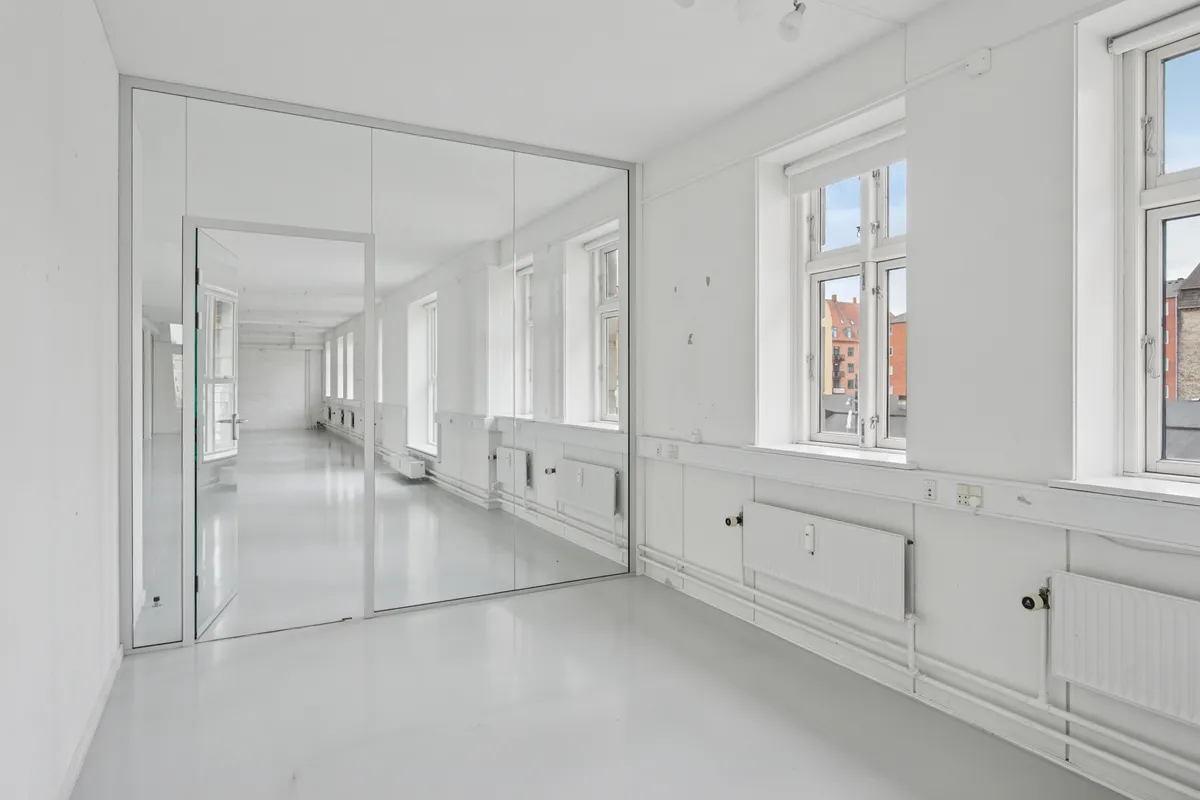 En lys og åben kontorry, med spejl og hvide vægge. Spejlet reflekterer rummet og giver det en ubegrænset følelse. Der er vinduer og radiatorer og lyst og rent gulv.