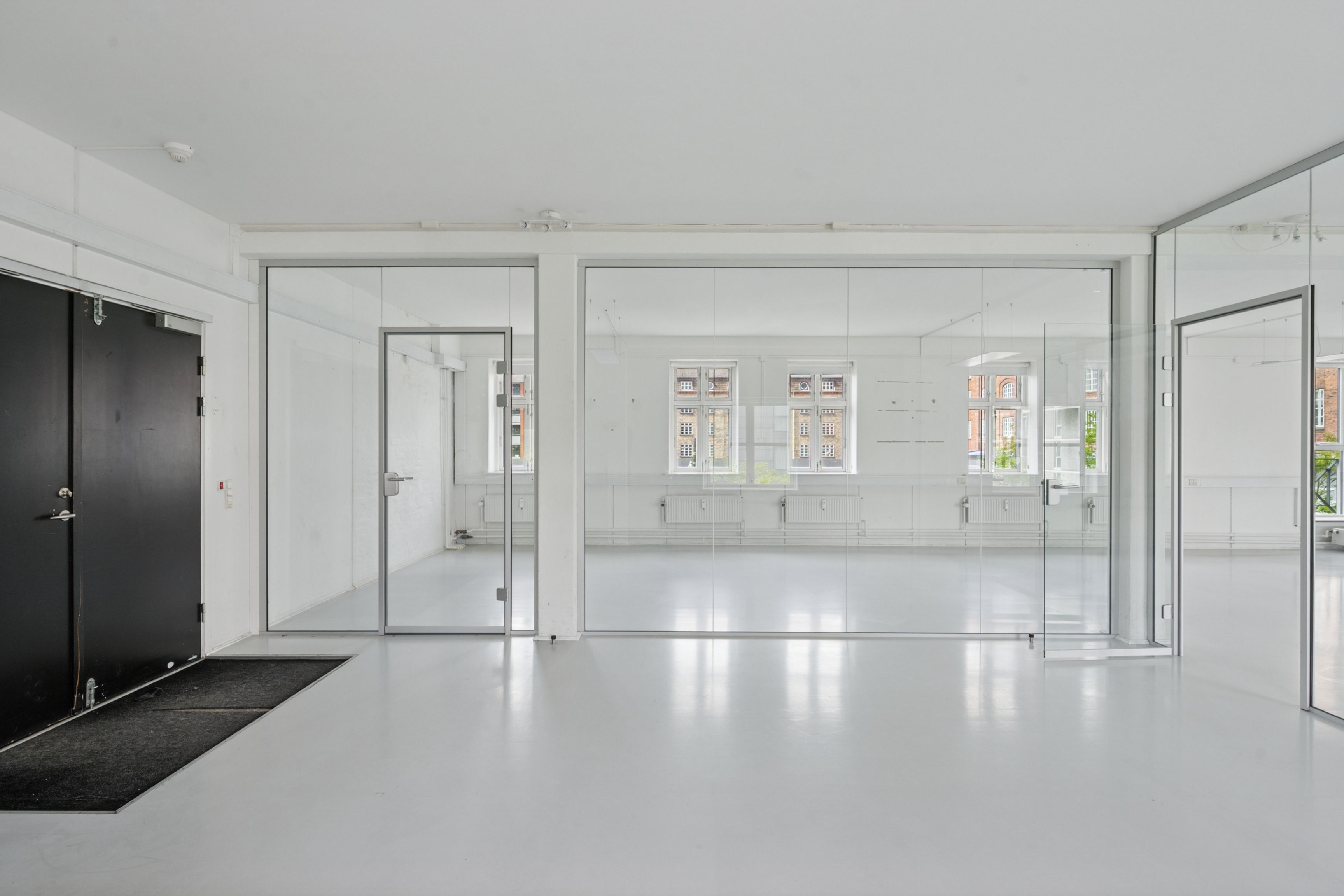 En stor kontorrum med hvide vægge og glasvægge samt lysegule gulve. En sort indgangsdør med gulvtæppe samt et glasparti med håndtag