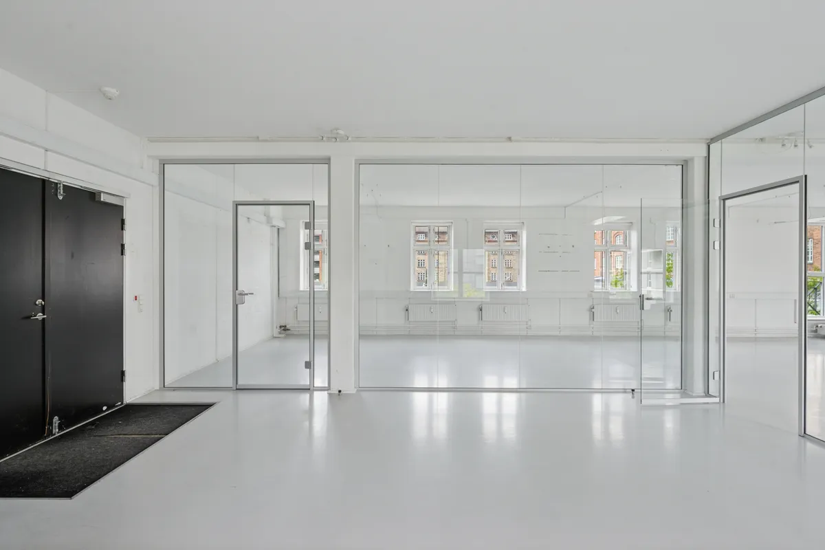 En stor kontorrum med hvide vægge og glasvægge samt lysegule gulve. En sort indgangsdør med gulvtæppe samt et glasparti med håndtag