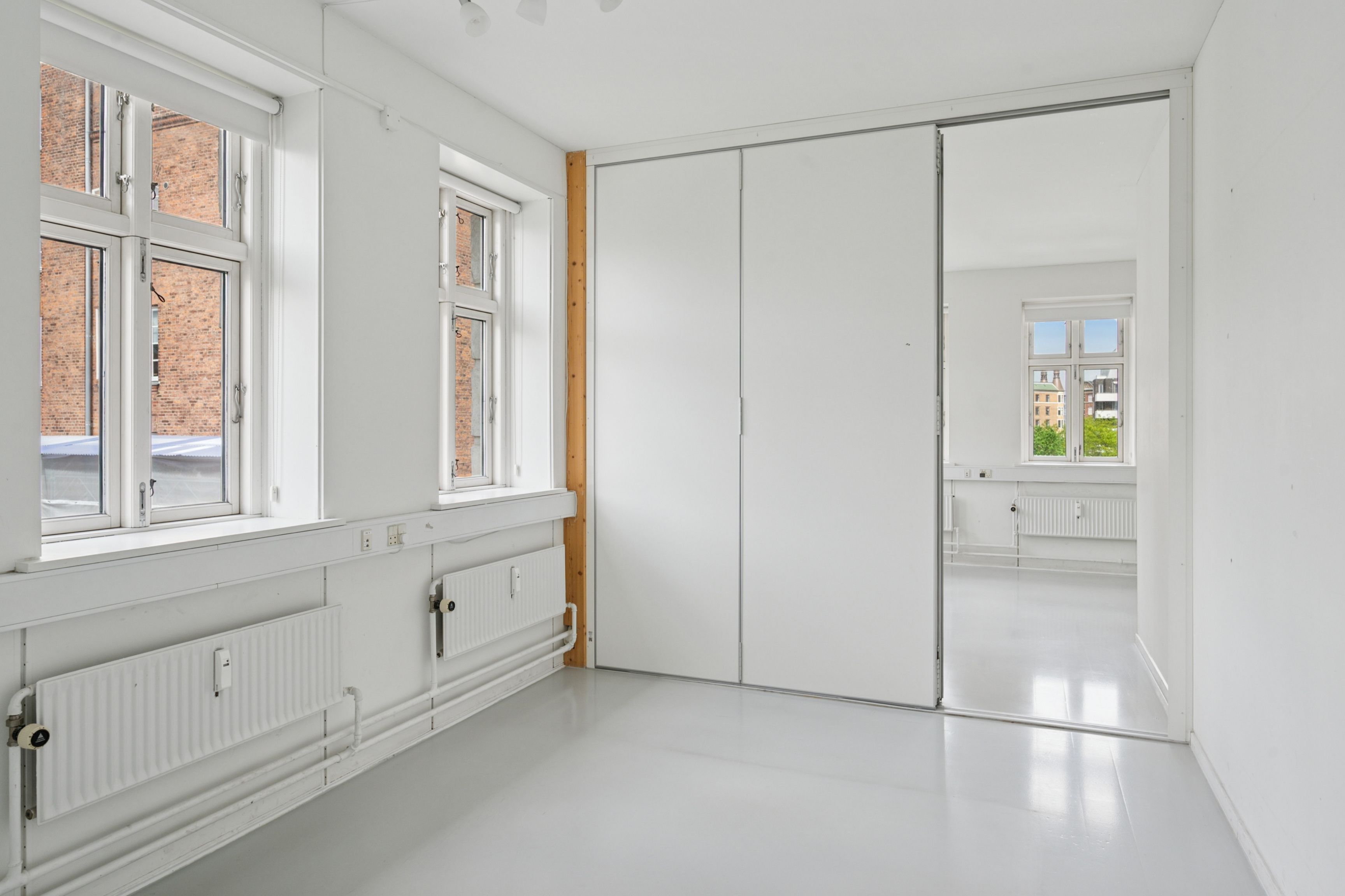 To rum med hvide vægge og højt til loftet, store vinduer og spejle, hvide gulvbelægning og radiatorer samt tre lysekroner i loftet