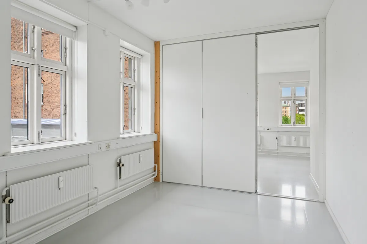 To rum med hvide vægge og højt til loftet, store vinduer og spejle, hvide gulvbelægning og radiatorer samt tre lysekroner i loftet