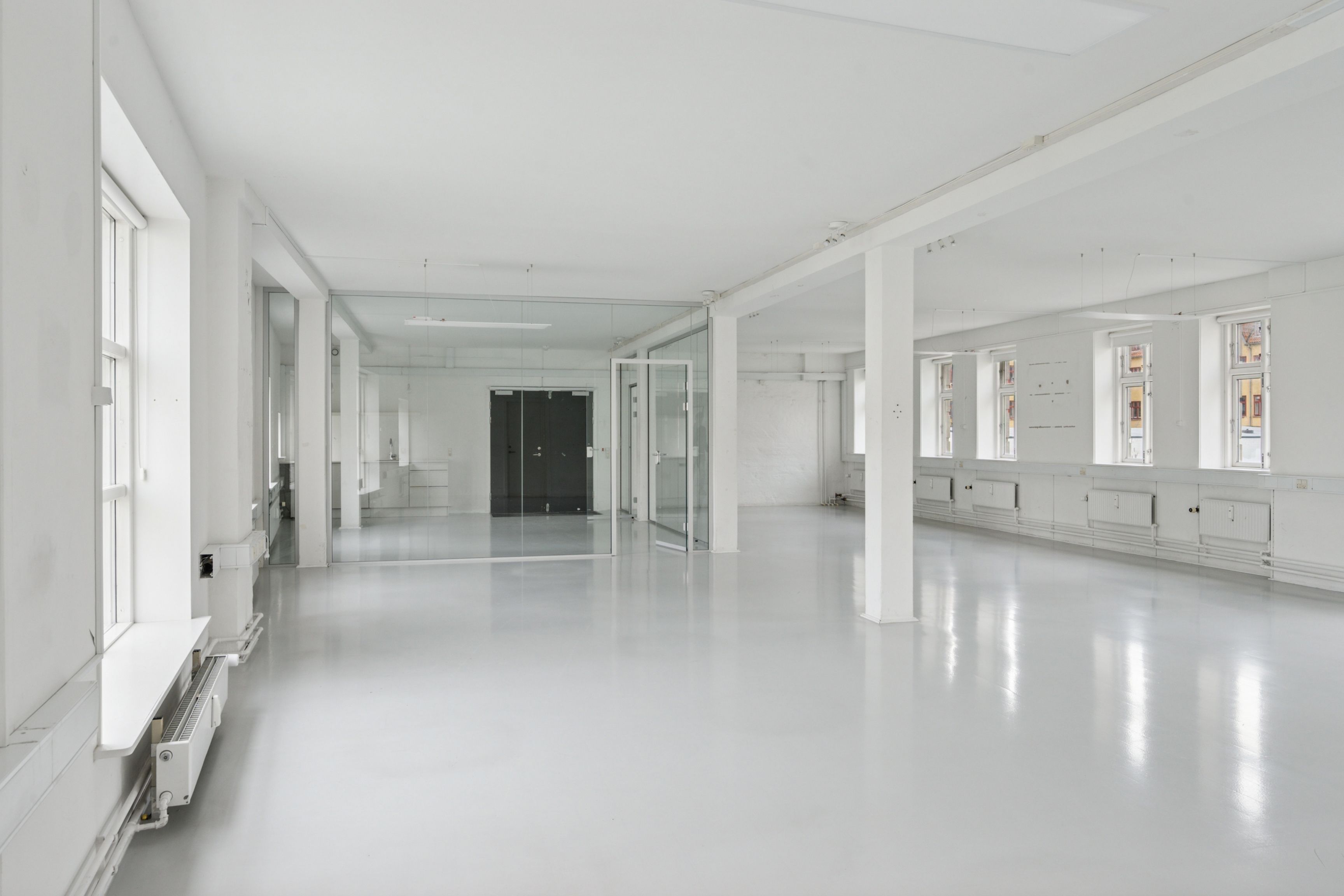 En stor og lys kontorlokal med hvide vægge og gulv og store glasvinduer og døre og hvide radiatorer og en lysekronehænger i loftet