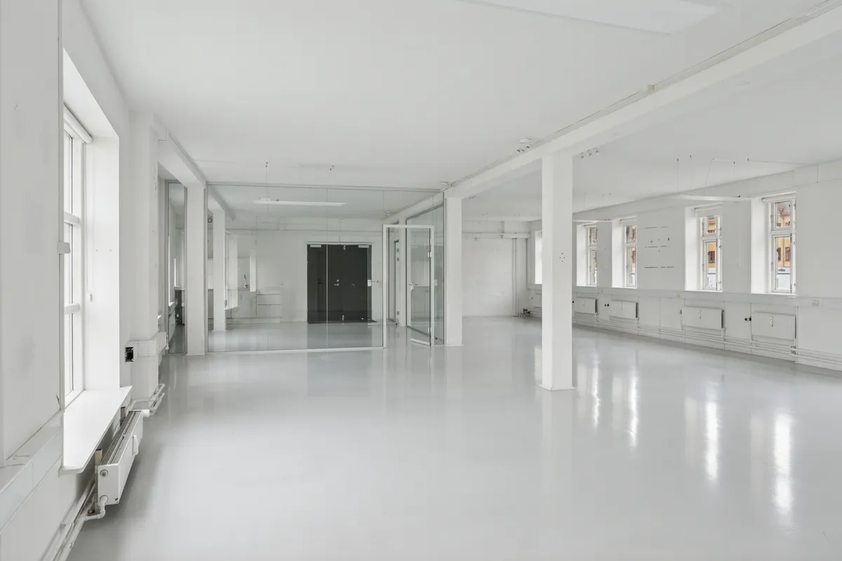 En stor og lys kontorlokal med hvide vægge og gulv og store glasvinduer og døre og hvide radiatorer og en lysekronehænger i loftet