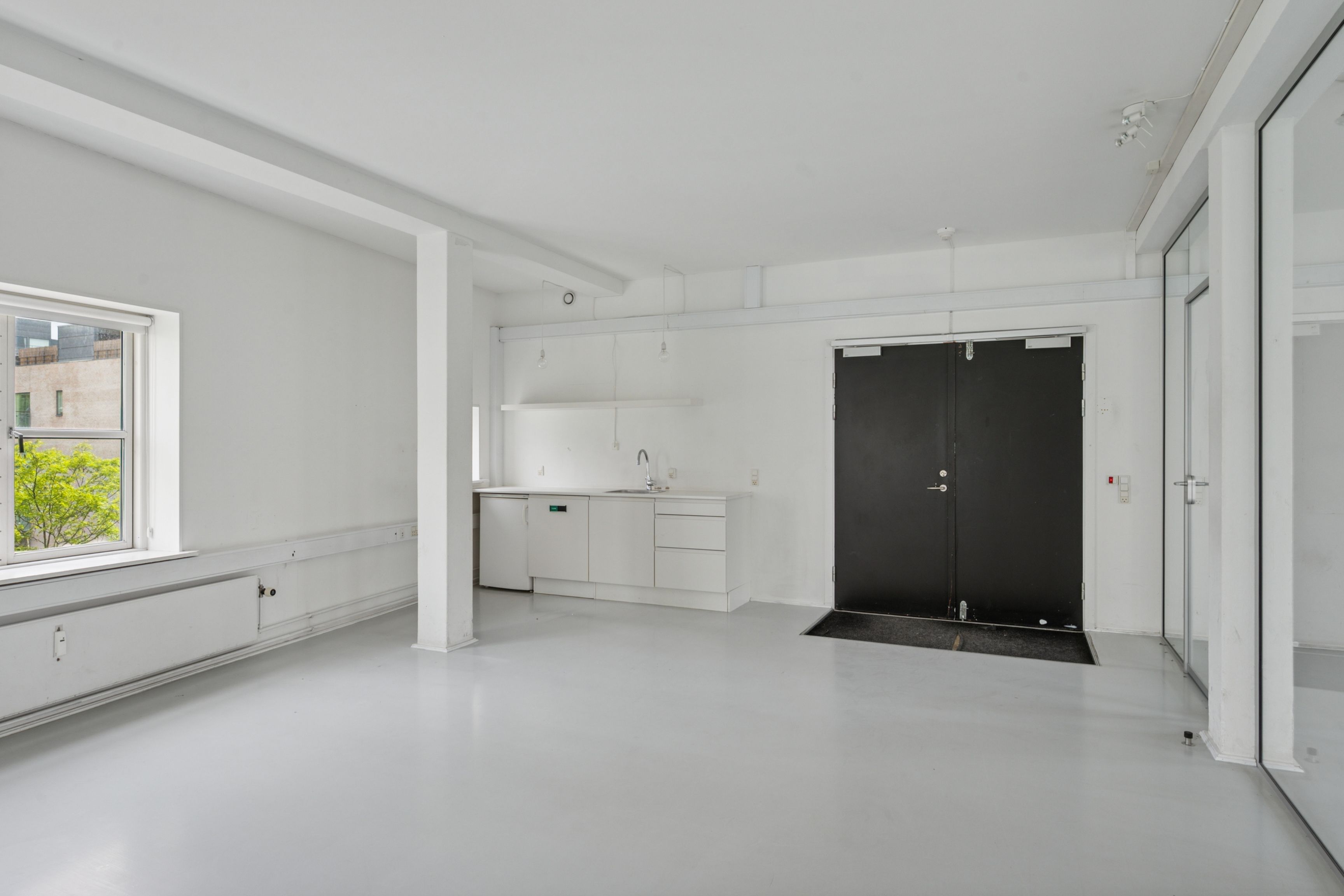 Lyst kontorlejemål med lyse lofter, hvide vægge og mørke døre. Indvendt køkken med vask og lysekroner. Vindue med udsigt til grønne træer og bygninger