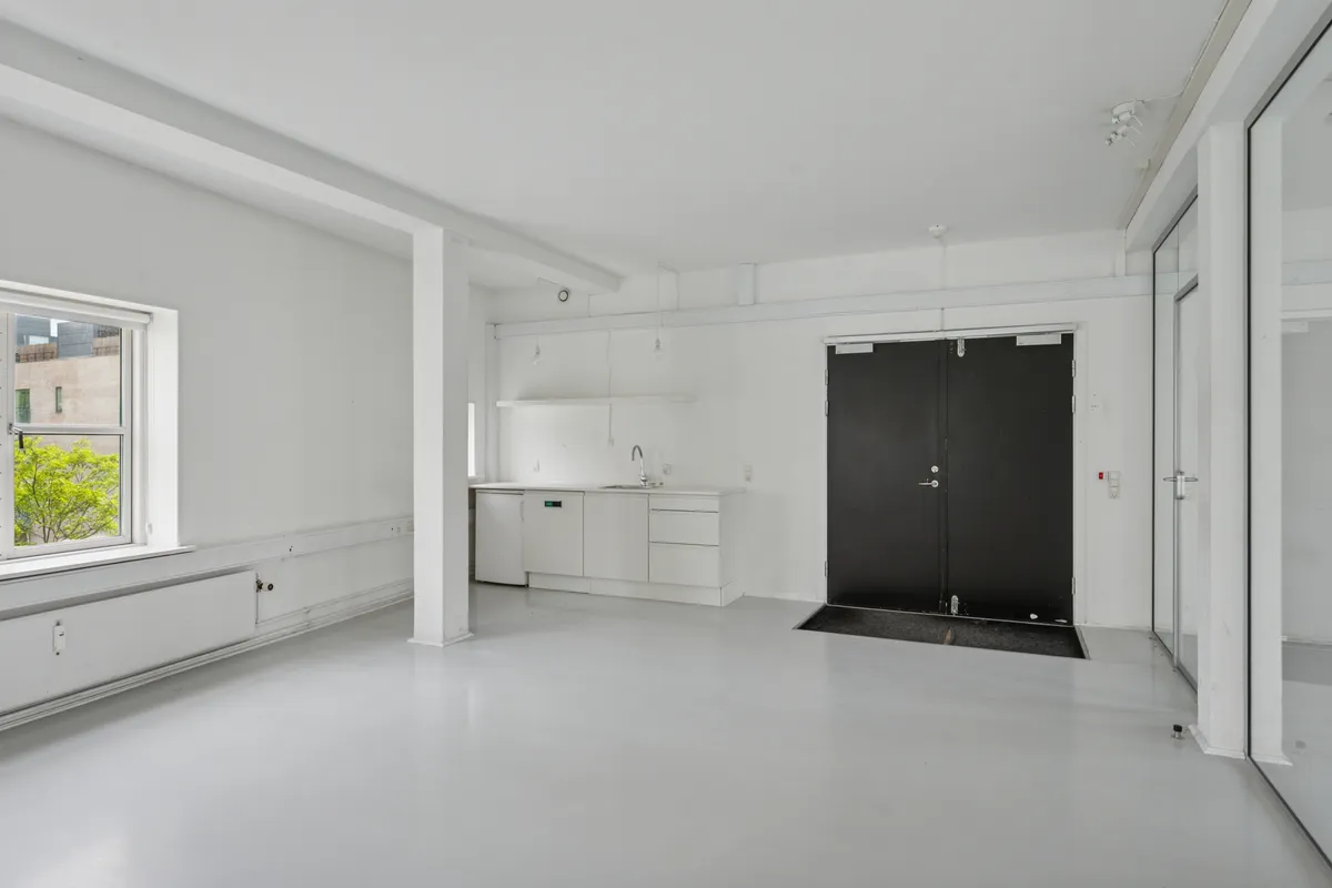 Lyst kontorlejemål med lyse lofter, hvide vægge og mørke døre. Indvendt køkken med vask og lysekroner. Vindue med udsigt til grønne træer og bygninger