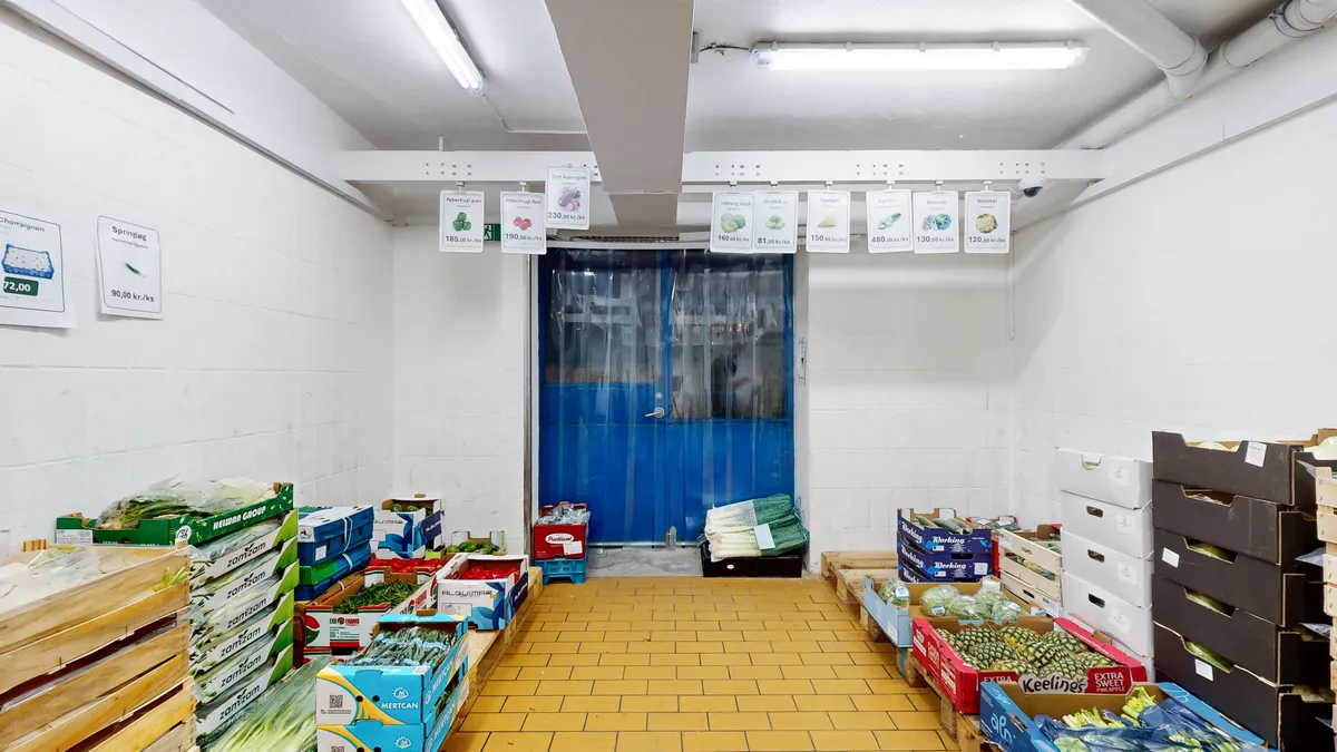 Lagerrum med gulvbelægning og lagerkasser med grøntsager, salat og andre frugter og grøntsager samt prisliste i en hvid og blå dør