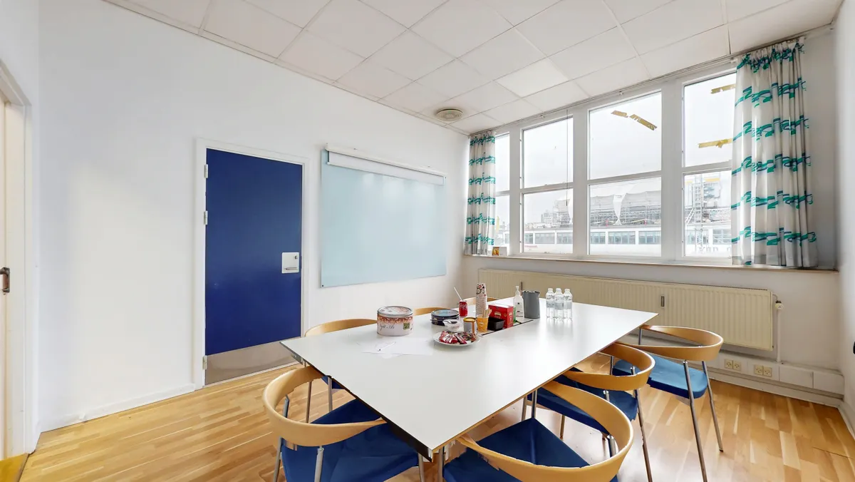 Hvidt mødelokale med ovalt bord og fem lysestole samt en dør og et vindue med udsigt over byen