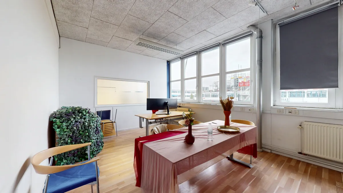 Store vinduer, en lang borde og to lyse loftslamper i en møderum med et trægulv og en grøn plante