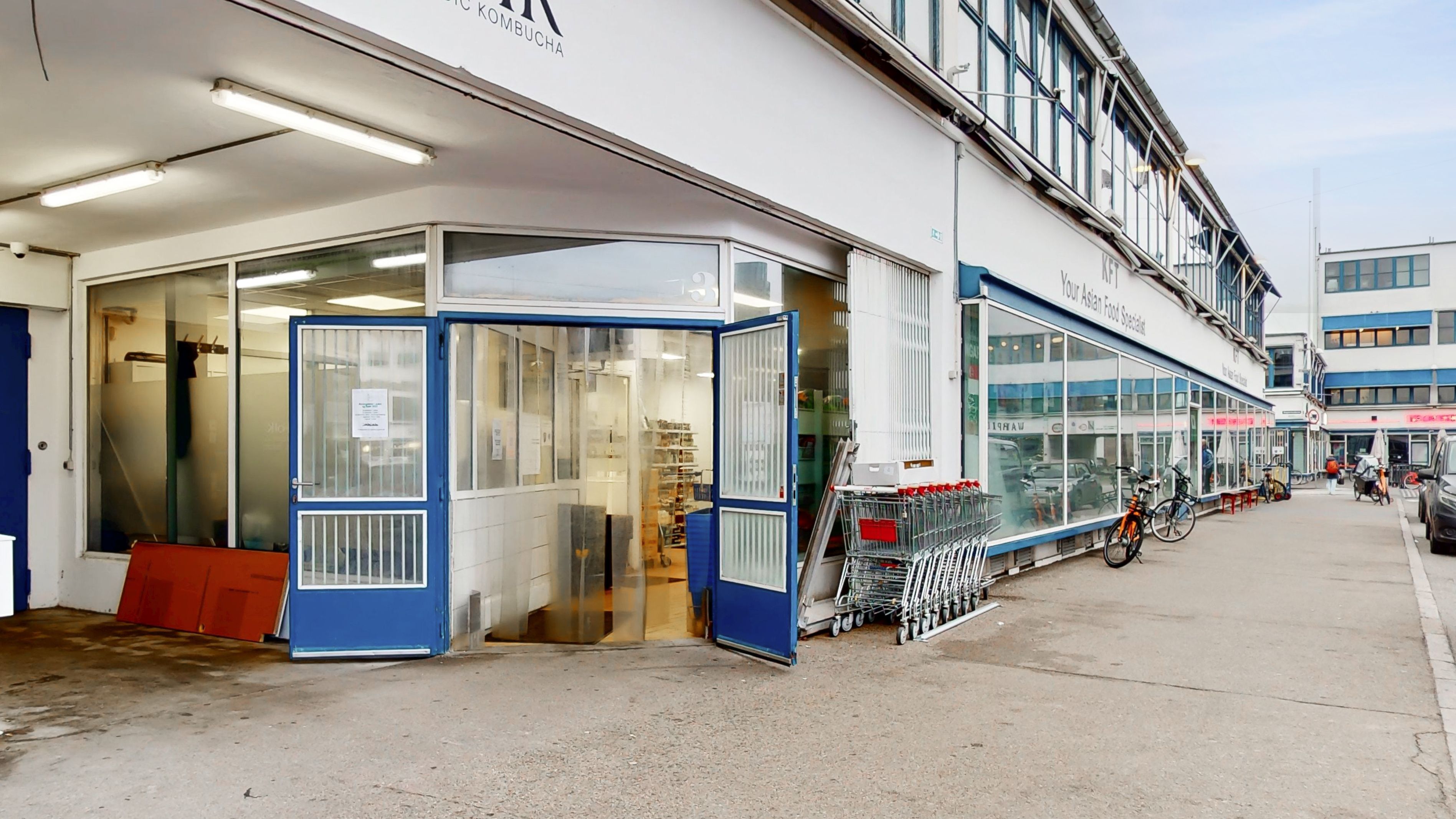 Store glasruder og åbne døre til erhvervslejemålet på Flæsketorvet. Parkerede cykler og indkøbsvogne udenfor. Lys udvendigt med skrift på facaden.