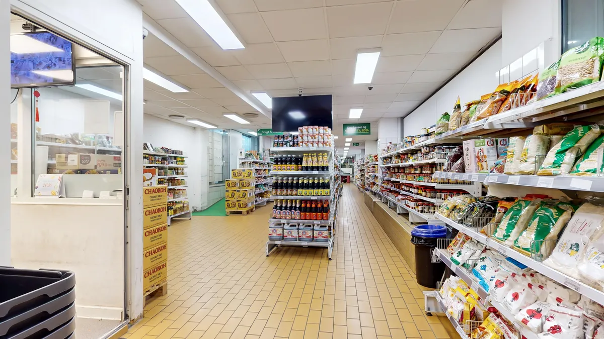 Lang korridor i supermarked med lysegulle fliser og hylder med fødevarer og drikkevarer. På hylderne er der flere forskellige varer, og der er et CCTV-kamera på loftet.