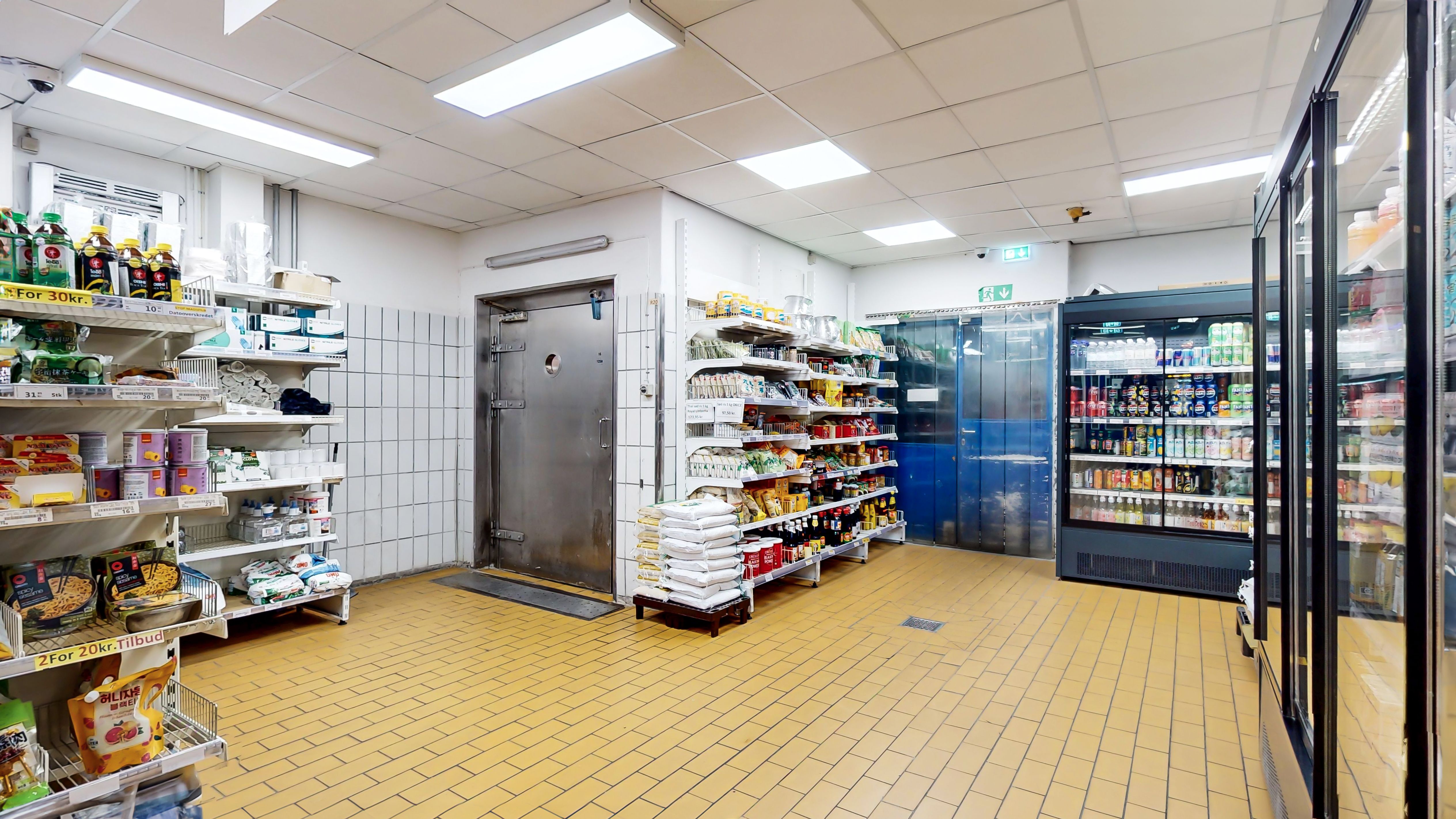 Supermarked med solgt og frosne fødevarer, gult belagt gulv og køleskabe med drikkevarer og snacks. En lagerdør er åben og et stokværk er i baggrunden