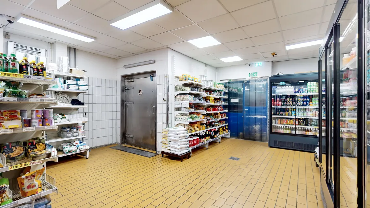 Supermarked med solgt og frosne fødevarer, gult belagt gulv og køleskabe med drikkevarer og snacks. En lagerdør er åben og et stokværk er i baggrunden