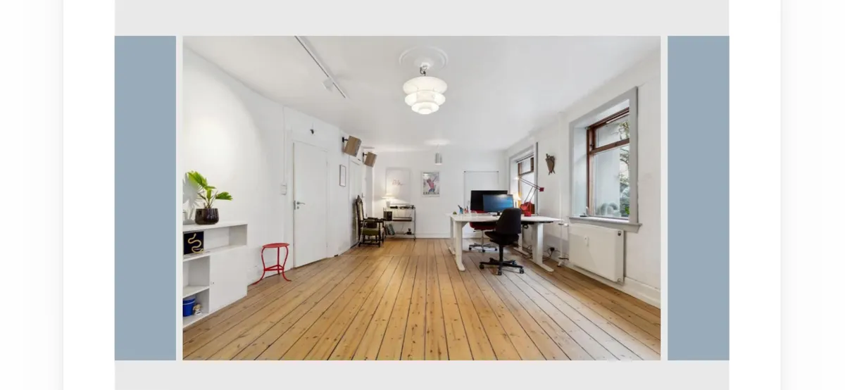 Inde i lokaler med højloft og lyse gulve. En plante og en stol står på højre side, og en bærbar computer er på bordet. Et sæt højtalere er placeret på gulvet.