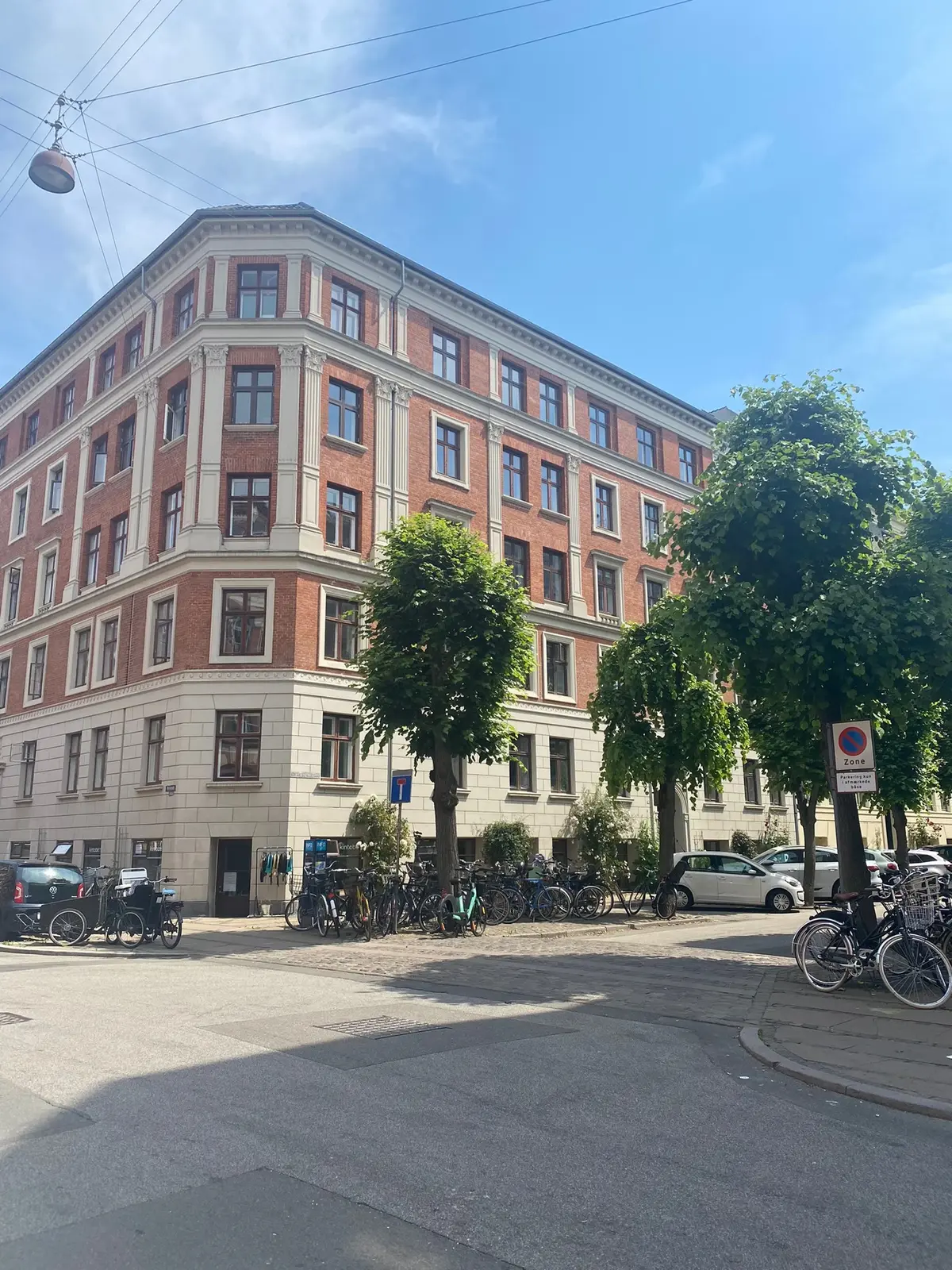 En stor, hvid og rødmaursten bygning med mange vinduer på Schleppegrellsgade. På forsiden står flere cykler og biler parkeret.