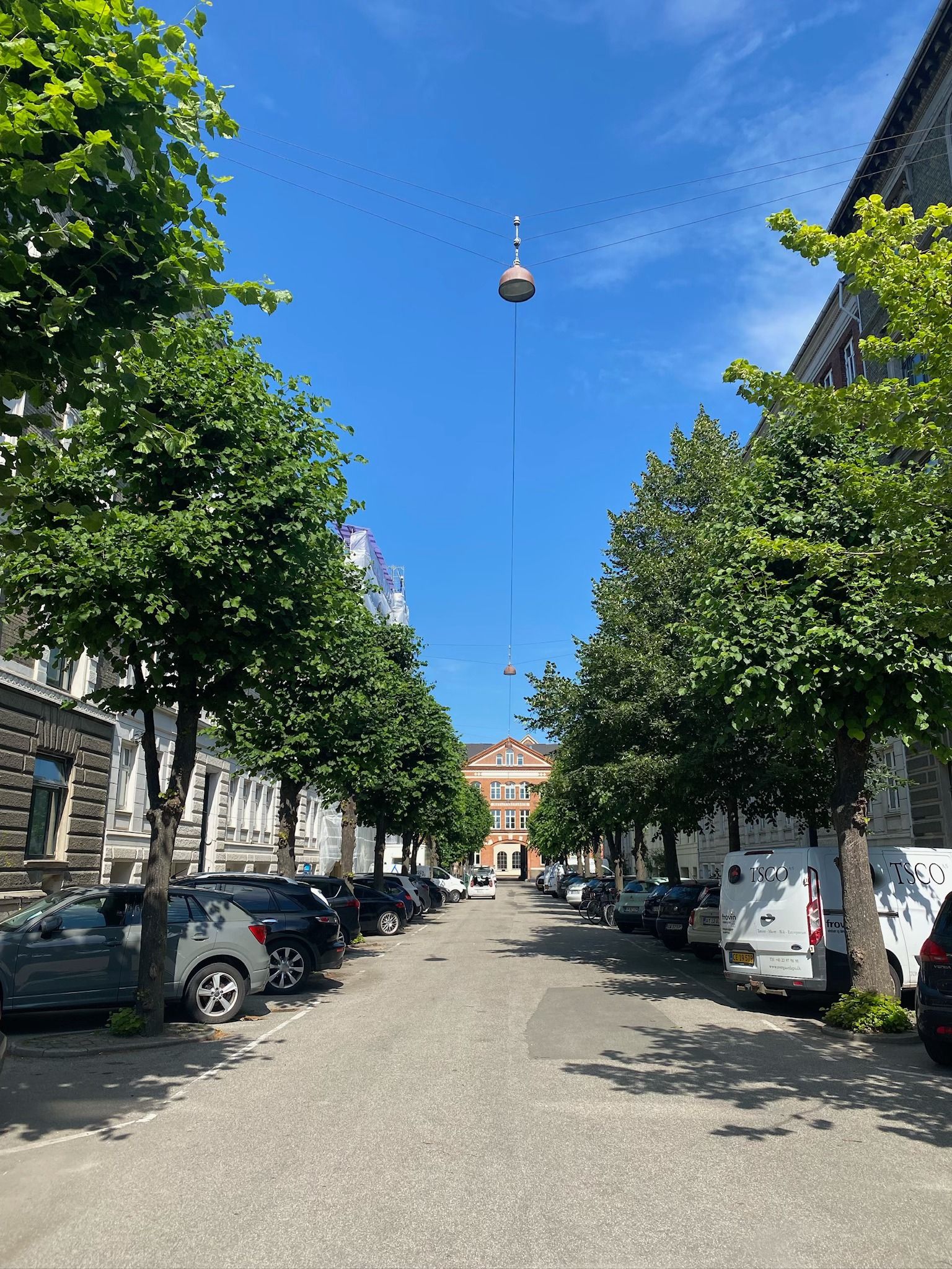 På Ryesgade ses parkerede biler og en træbevoksning. En vandret stang stiger op mod himlen, hvorfra en stang er hængt. Bygninger er opført langs gaden