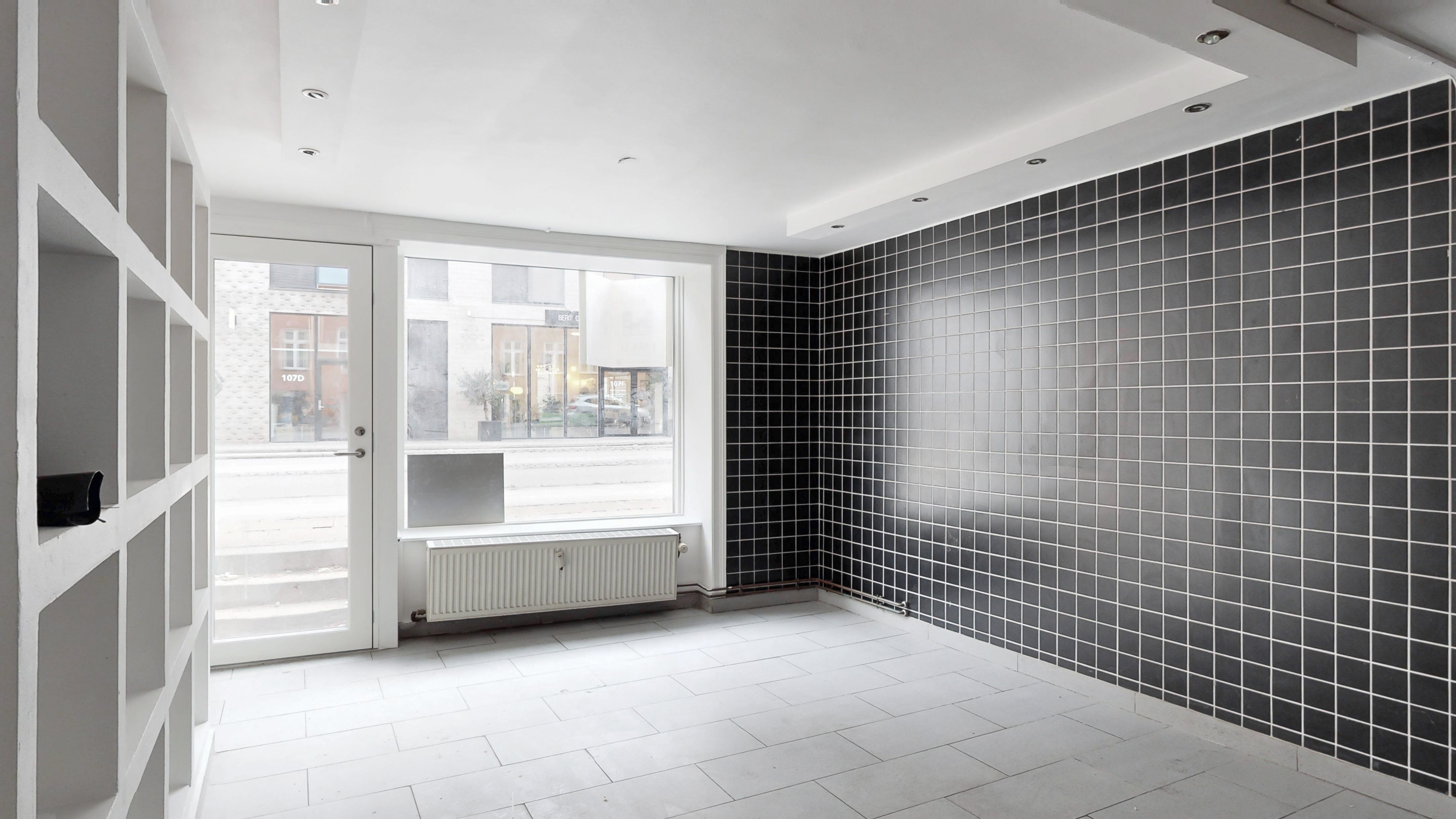 Minimalistisk kælderlejemål med hvide vægge og sort kakelbeklædning, radiator og glasdøre, der fører ud til Vesterbrogade.