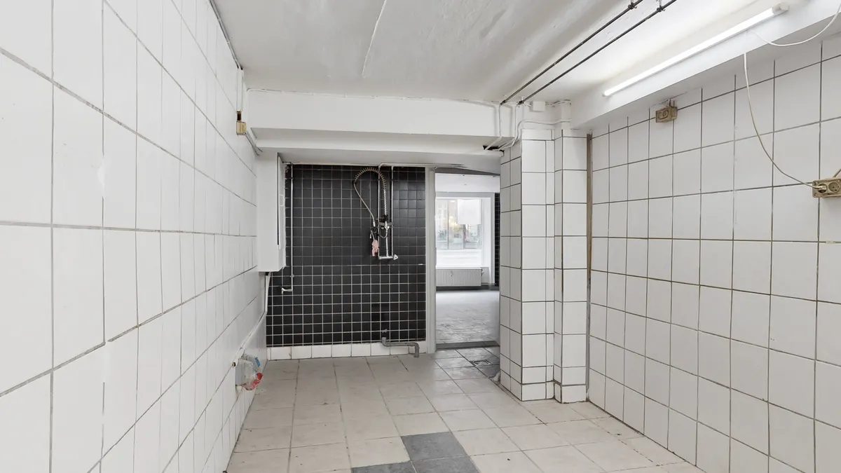 Kort gang i kælderlejemål med hvidt gulv og vægge, tørrehylde og røret med slange i mørkt badeværelse.