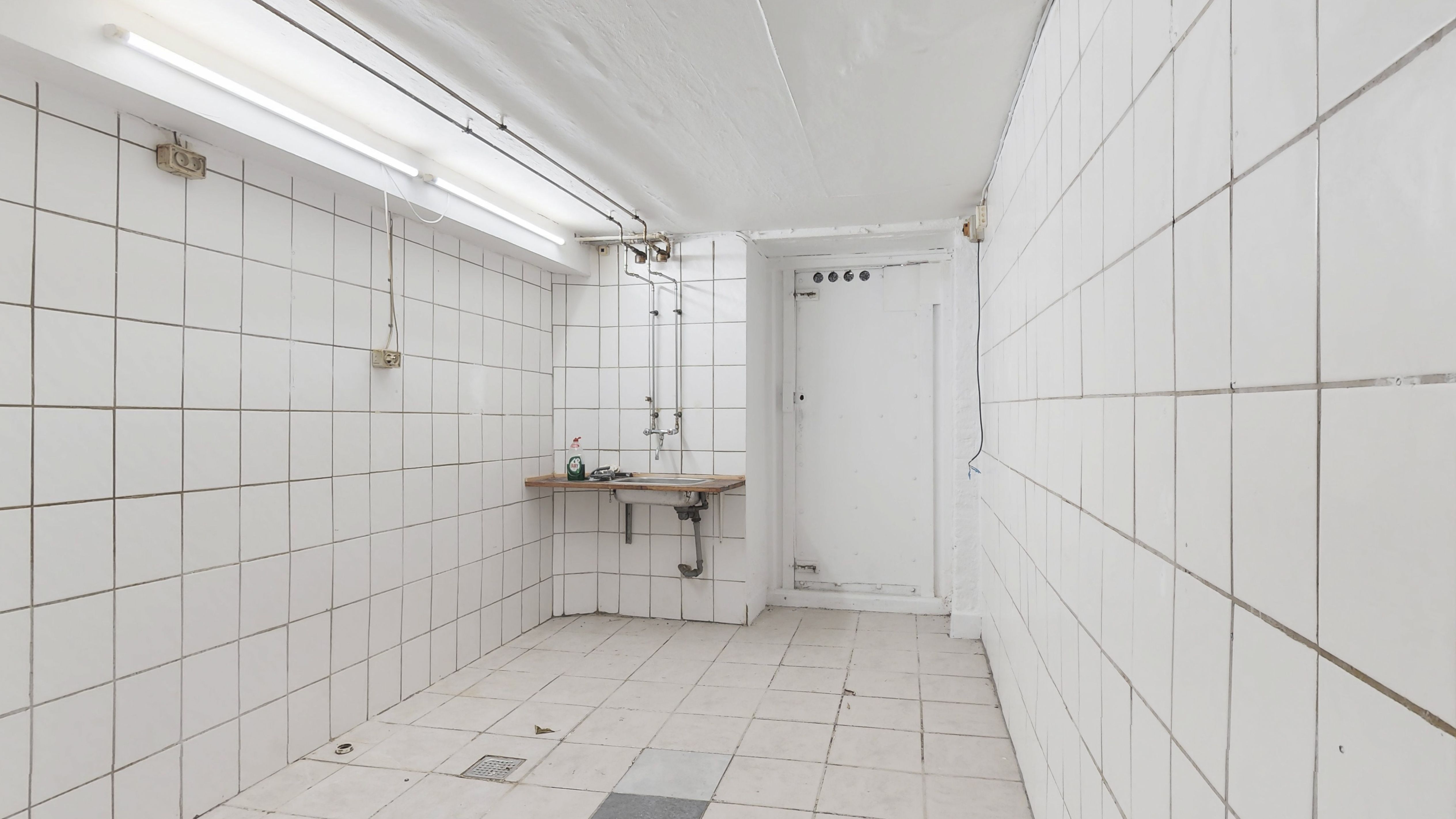 En hvidt kledet kælderrum med en lille vask og tøjvasker og et håndklæde. Lysestage på loftet og hvidt flisegulv.