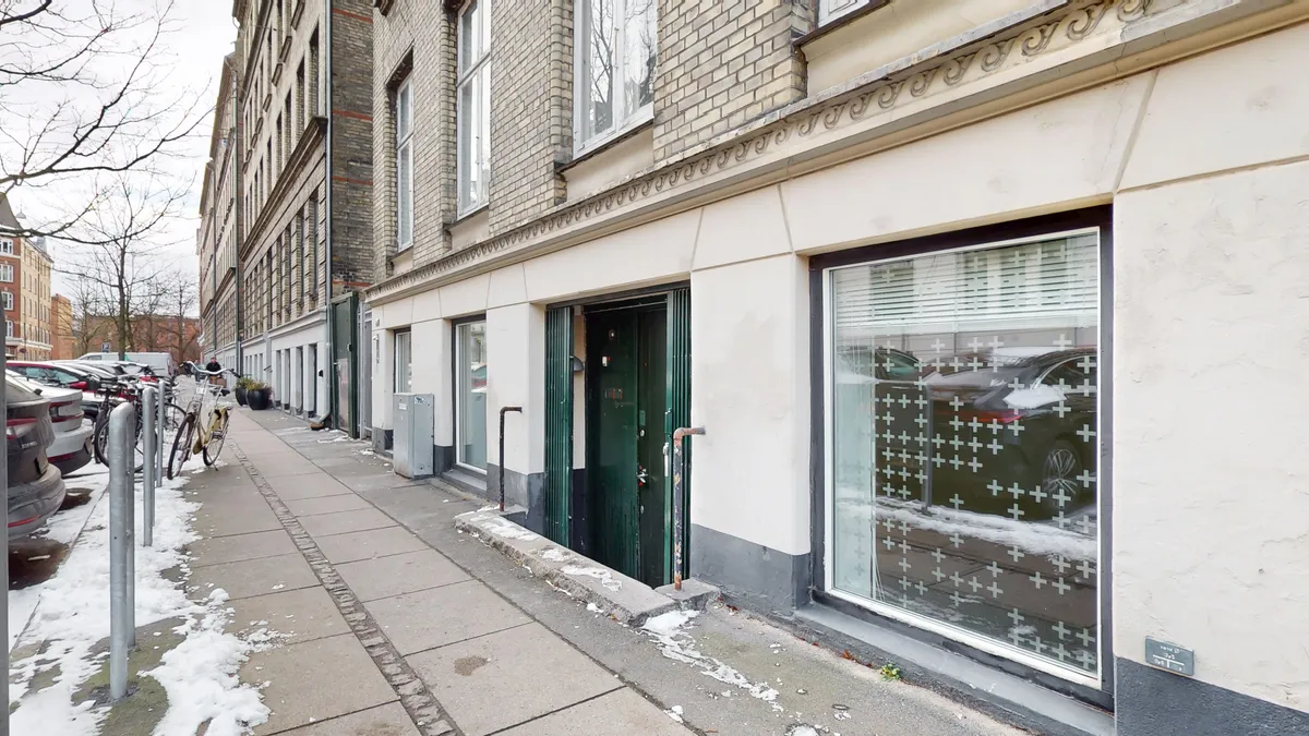 Forretningslejlighed med grønne døre og sne på sidevæggen og gulvet. Parkerede biler og en cykel med kurv på forsiden.