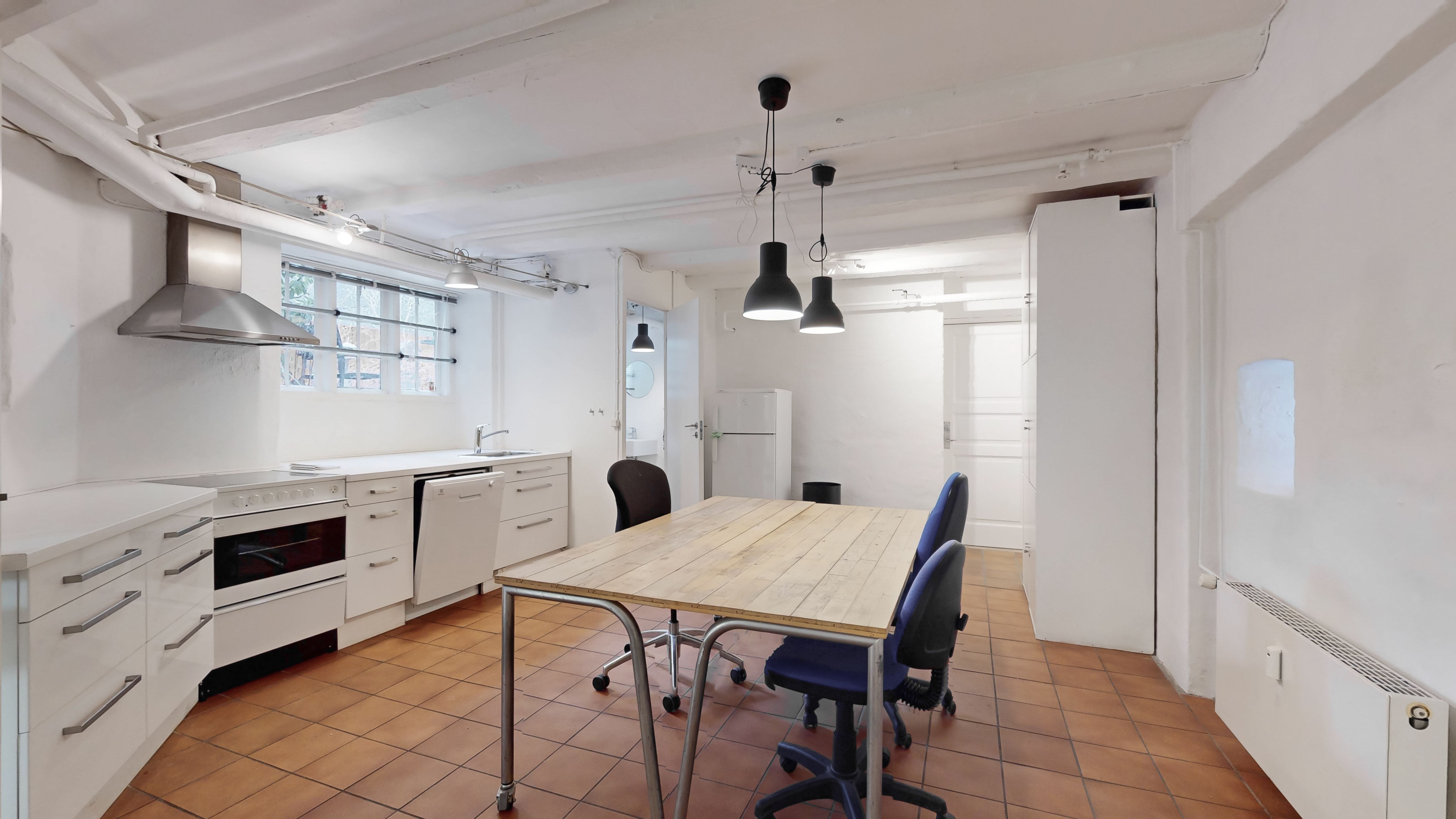 Et enkeltværelse med moderne køkken og lyse loftslampe, hvidt køkken med ovn og vaskemaskine samt to lyse stol ved et bord