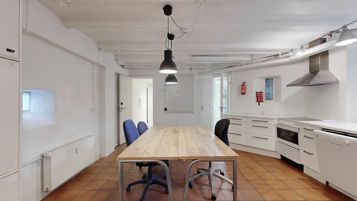 Værelse med højt til loftet, tre lysekroner, tre stole, bord, køkken med ovn og komfur, lysekroner og en rød ildslukker