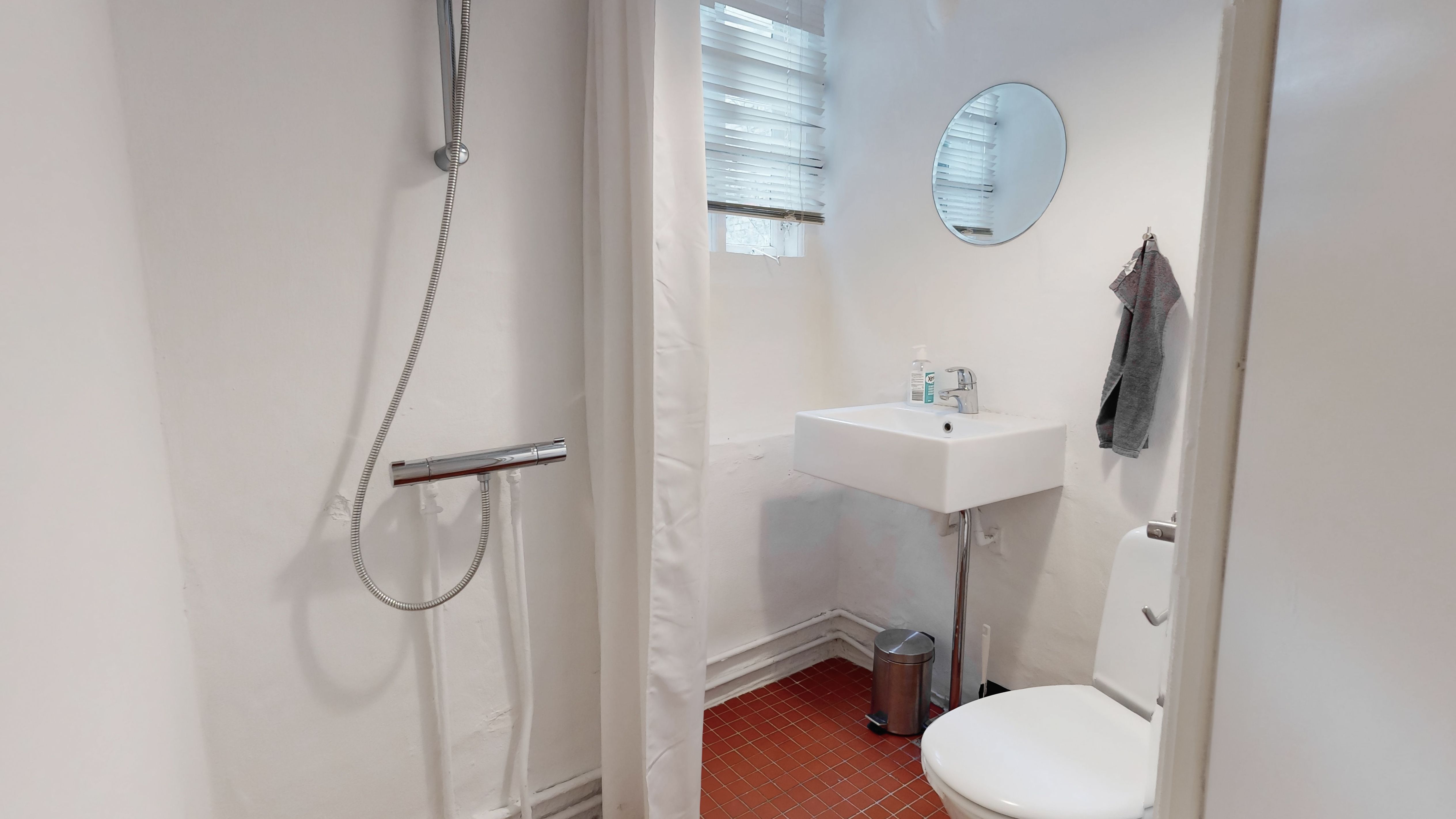 Et lille badeværelse med toilet, hvidt badekar og en rund spejl på væggen. Et håndklæde hænger på hængekrogen og der er en tøjskaft i badeværelset