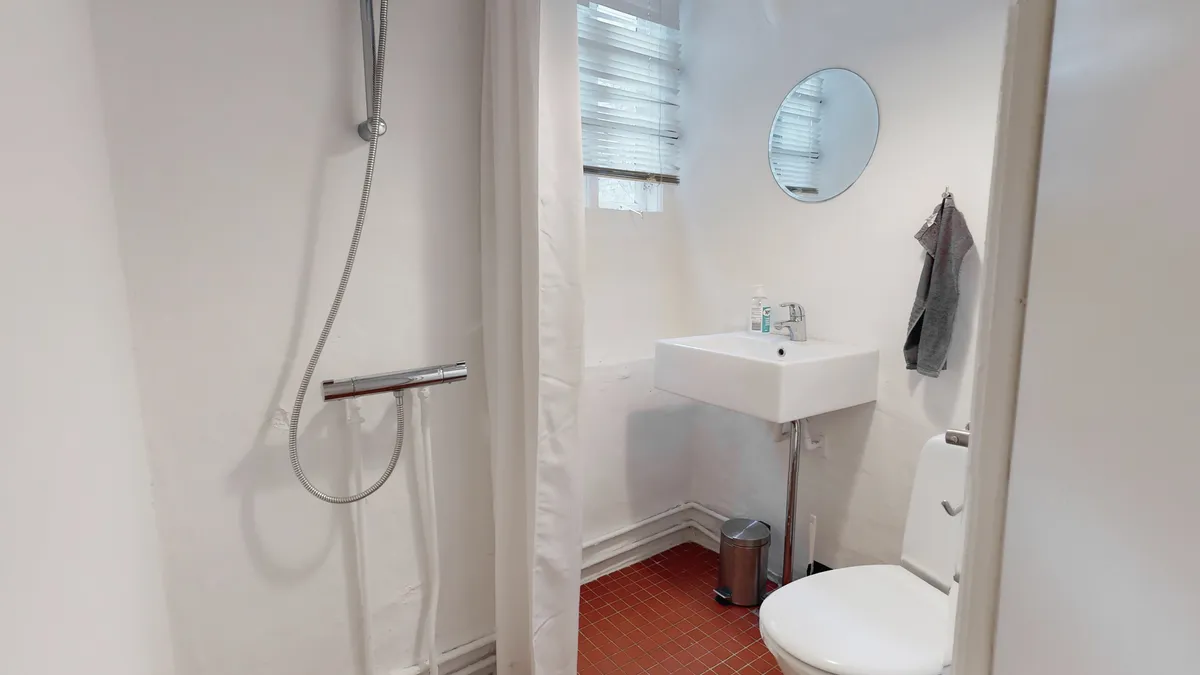 Et lille badeværelse med toilet, hvidt badekar og en rund spejl på væggen. Et håndklæde hænger på hængekrogen og der er en tøjskaft i badeværelset