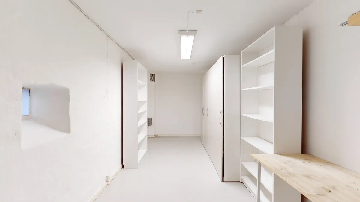 Langt, ubenyttet værelse med lyse loftslys og hvide vægge og gulv samt høje hvide hylder og et træbord til højre.