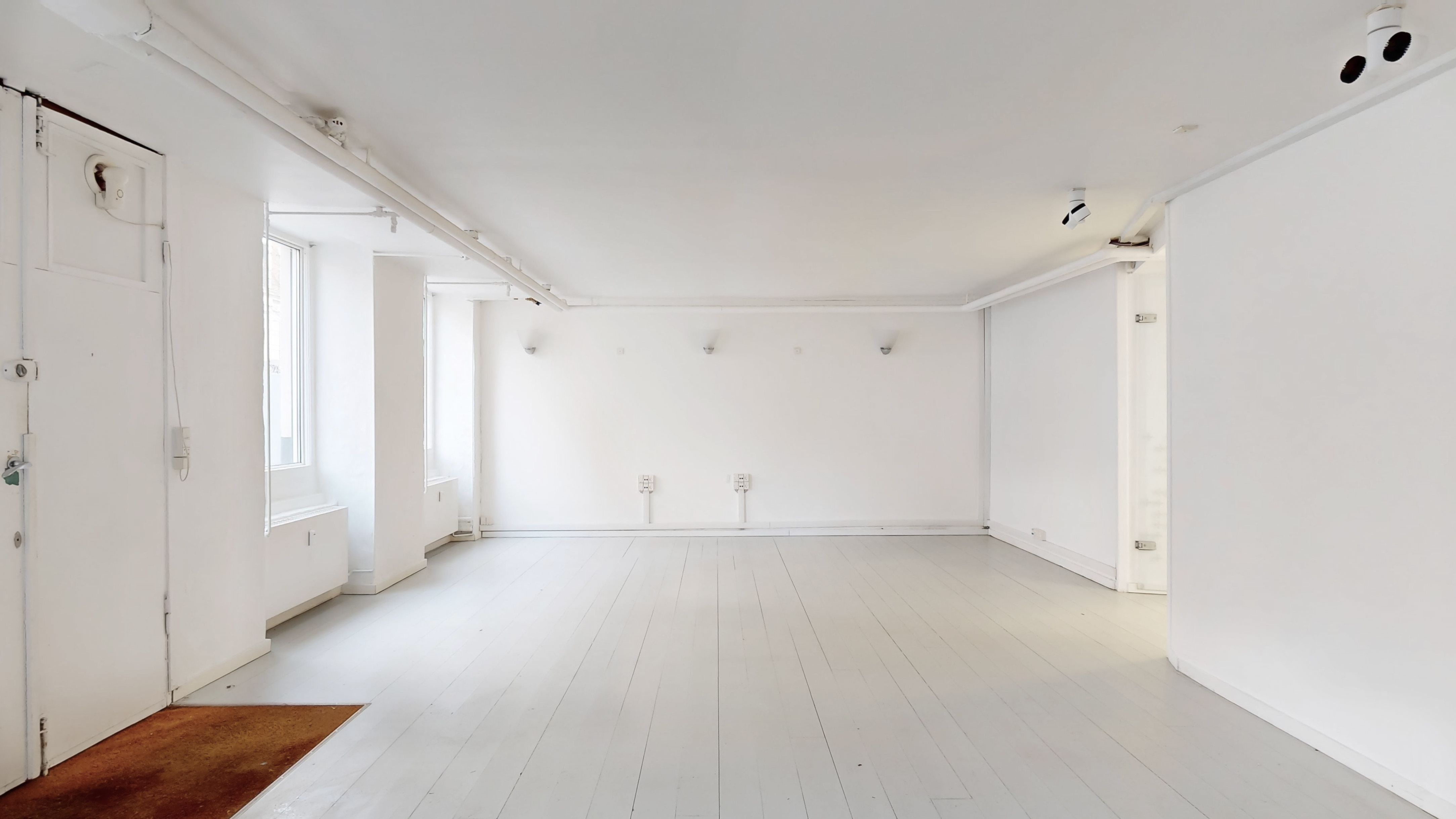 Store, hvide vægge og gulve i en tom erhvervslejlighed med fire lysekroner og et kamera. To vinduer giver udsigt til en sidegade.