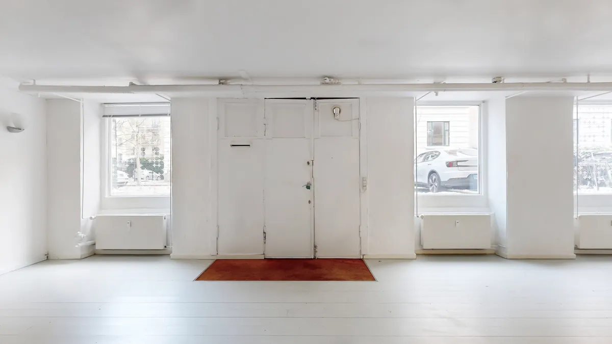 Erhvervslejlighed med hvide vægge, moderne indretning, og lyse vinduestage med udsigt til biler og gadekøer i baggrunden