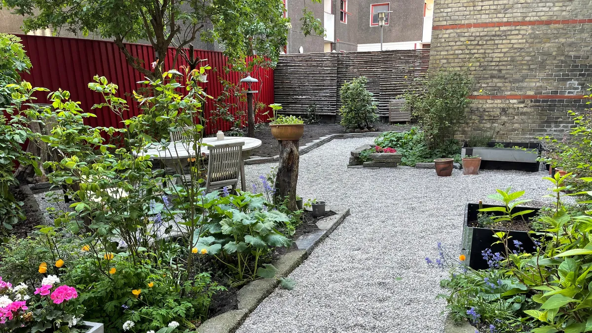 Have med små blomster, planter og lyserøde tulipaner, samt udendørs møbler og træværk, der stiger op mod en bygning med røde vindueskarme