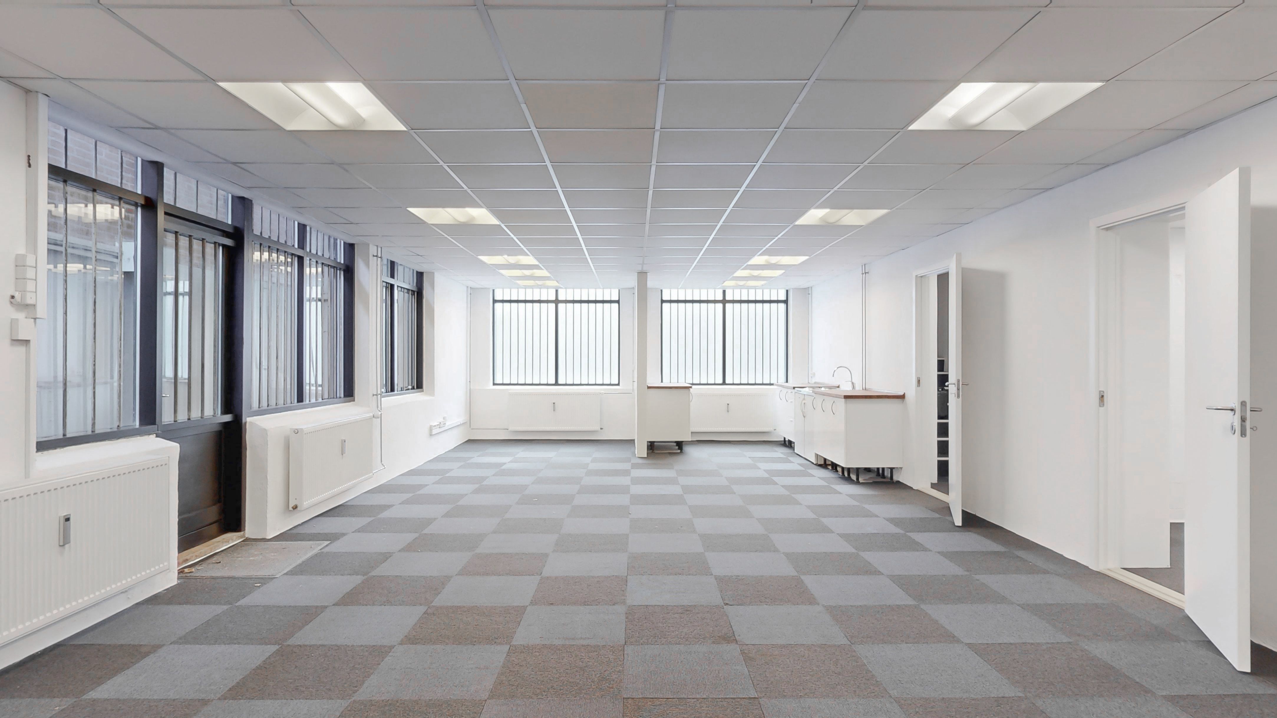 Store rummelige lokaler med hvide vægge, grå og hvide krydsvise gulvbelægning, radiatorer, hvide køkkenskabe og vinduer med stænger og ruder