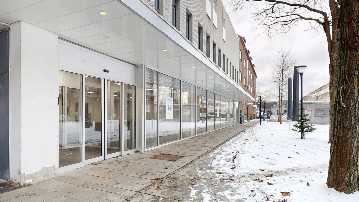 Butiksfront i hvidt og glas med indgangsdør og rektangulært vindue i Birkerød. Snedekoration og snedækket udenfor og en gågade med søjler og en fyr