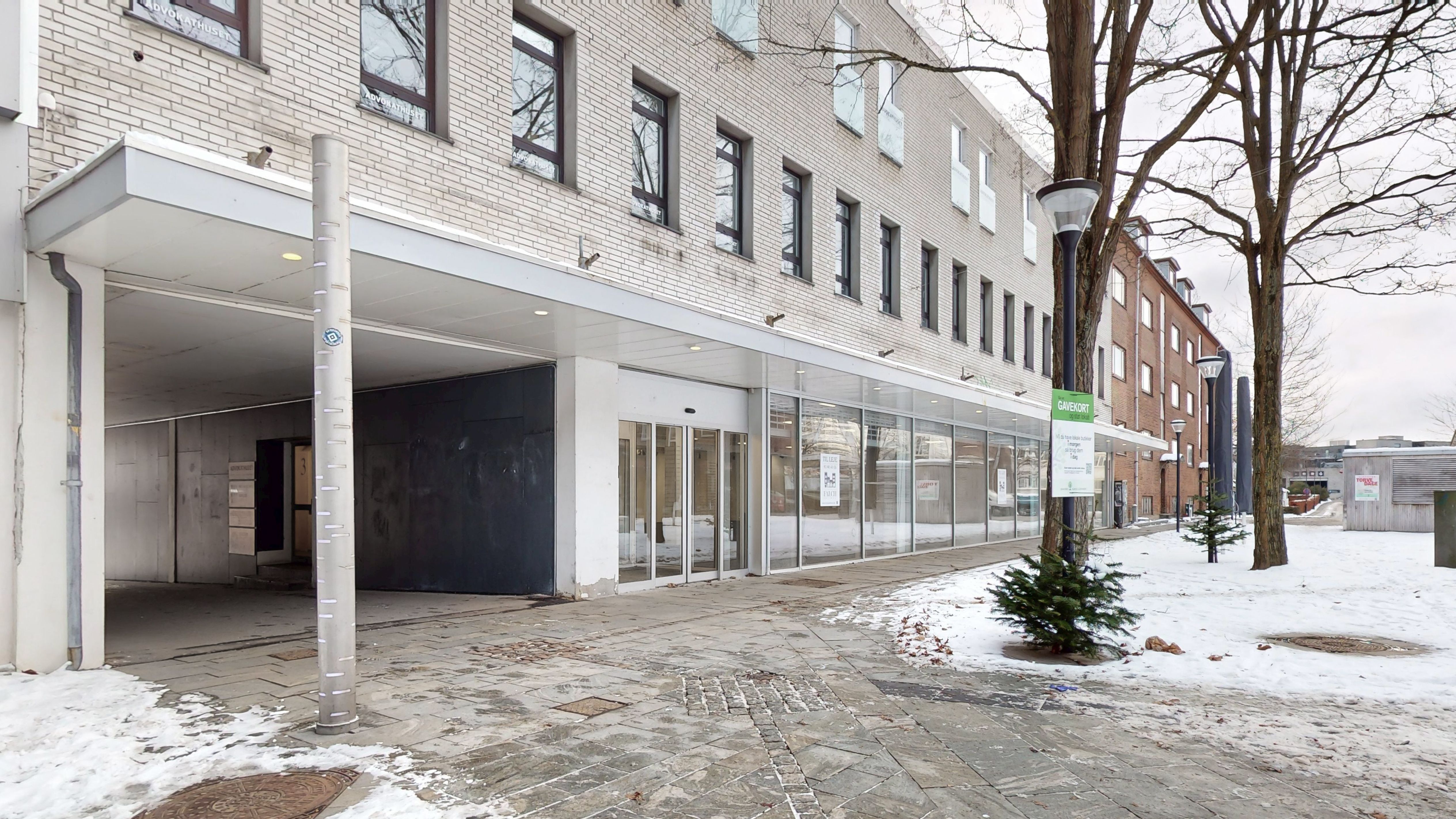 En moderne bygning med store vinduer og skilte, hvor en gade er dækket af sne med træer og et skovklædt område i baggrunden