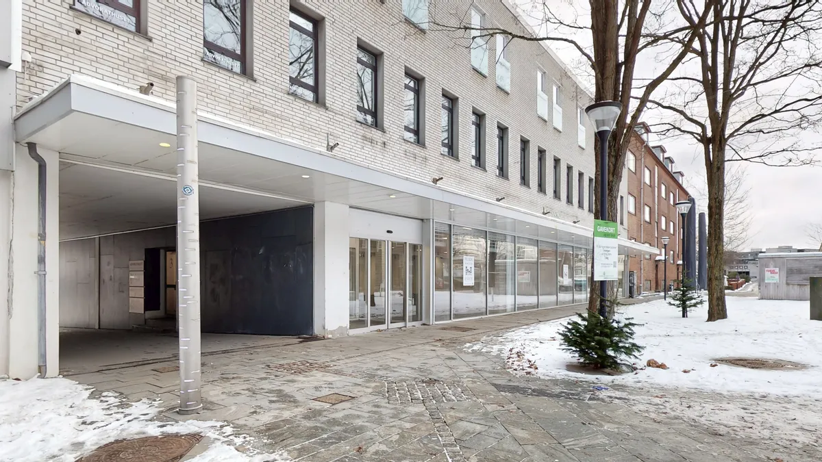 En moderne bygning med store vinduer og skilte, hvor en gade er dækket af sne med træer og et skovklædt område i baggrunden