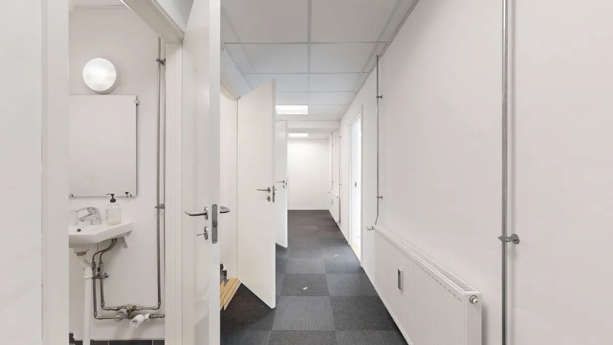 En lang, tæt lukket gang med hvide vægge og gulv, to åbne døre, hvor en er en toiletdør med spejl og håndvask, og højre væg med radiatorer og rør.