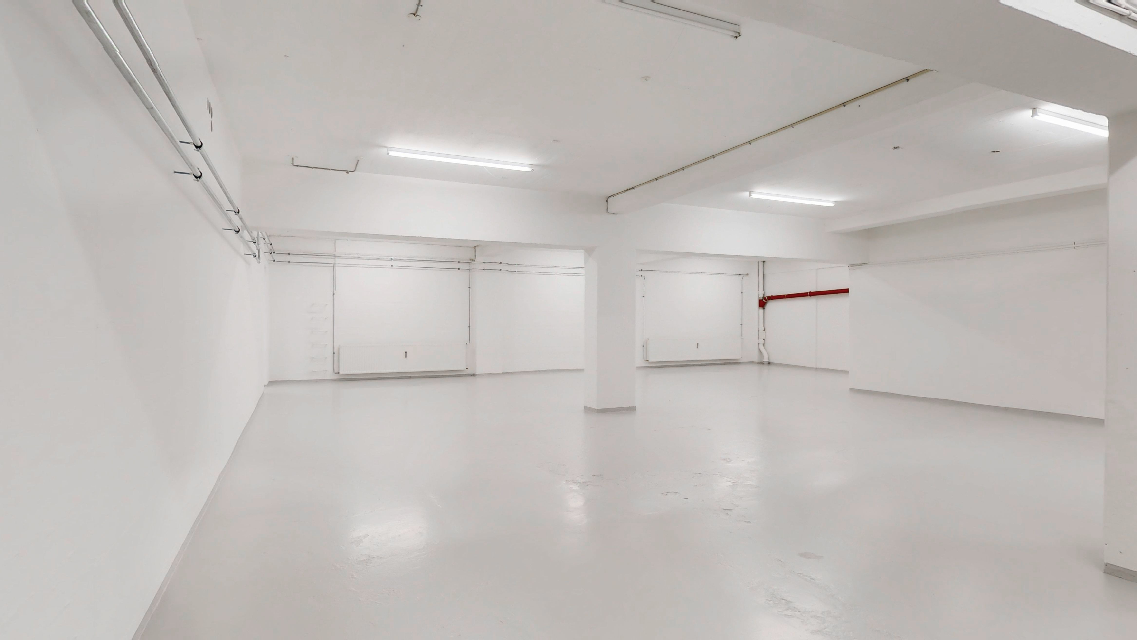 Et velbeliggende erhvervslejemål med højt til loftet, to store garagerum, og et stort lagerrum i kælderen. Gulvet er hvidt og blankt, og der er ingen møbler i rummet
