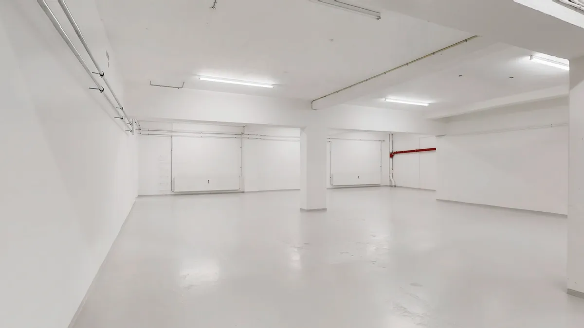 Et velbeliggende erhvervslejemål med højt til loftet, to store garagerum, og et stort lagerrum i kælderen. Gulvet er hvidt og blankt, og der er ingen møbler i rummet