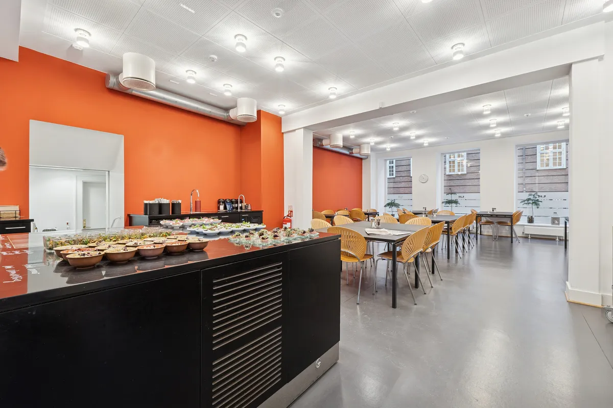 Kontorlokaler med moderne møbler, hvide vægge og orange accenter i lokalerne, fuld af serveringsborde med mad og drikkevarer samt lysekroner