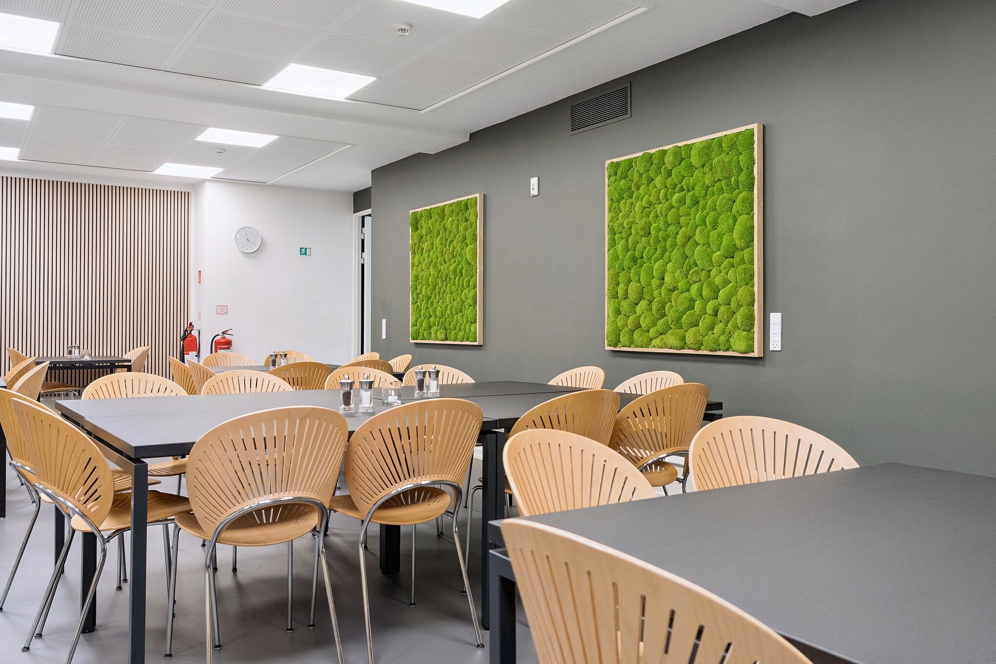 Kontor med lyse lofter og grønne vægdekorationer, møbler og borde med stole og en klokes klokker og ildslukker i baggrunden