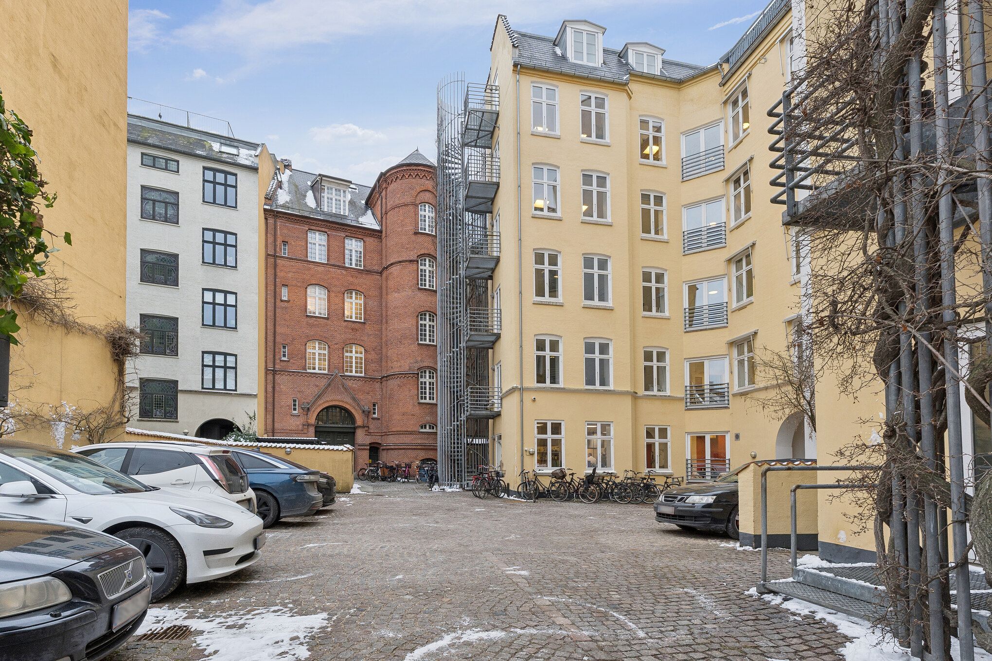 Flerbrugerhus med parkeringsplads, kontorlokaler og parkerede biler i København. Kontorbyggeri med moderne design og lyse vinduer. Forskelligt farvede bygninger med buske og snedækket.