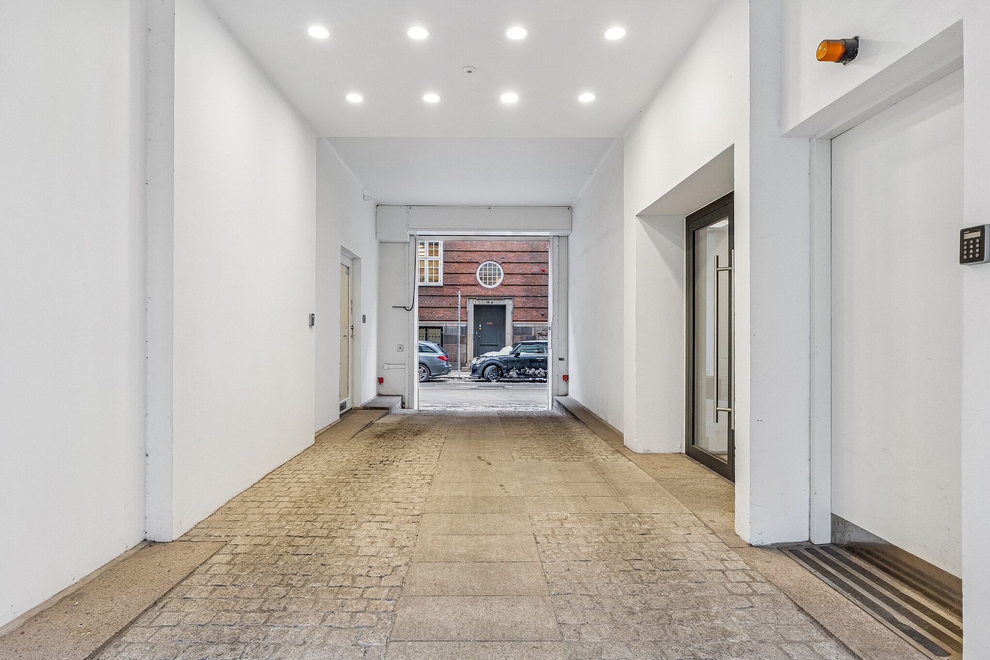 En moderne, lys og rummelig gangvej i et flerbrugerhus med en række biler parkeret udenfor og en brun bygning i baggrunden