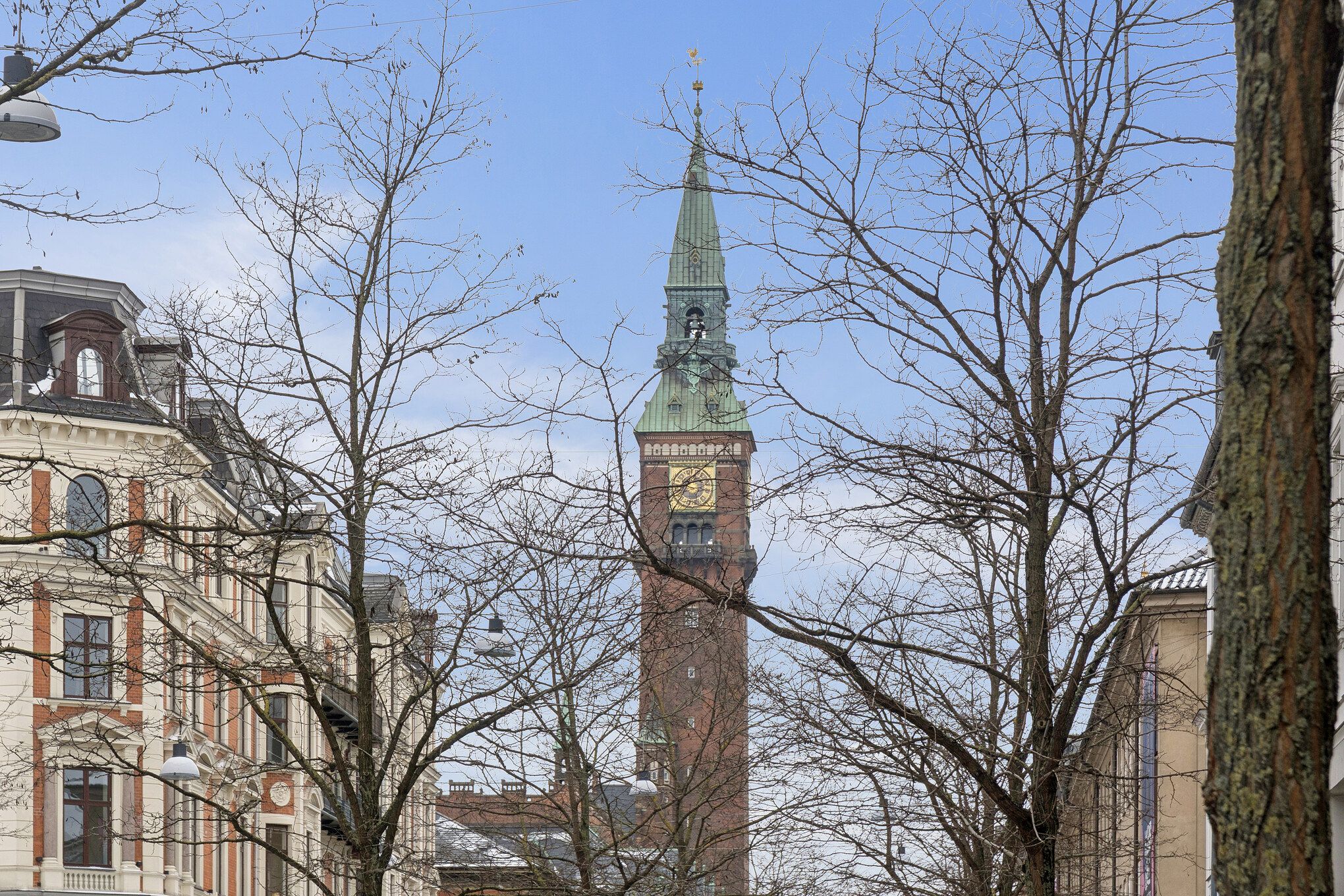 Talrige gamle bygninger og træer med lyseblå himmel. Et støbejernslys og en klokke på en høj slank tårn. En gul urtavle med sorte tal og skrift på tårnet.