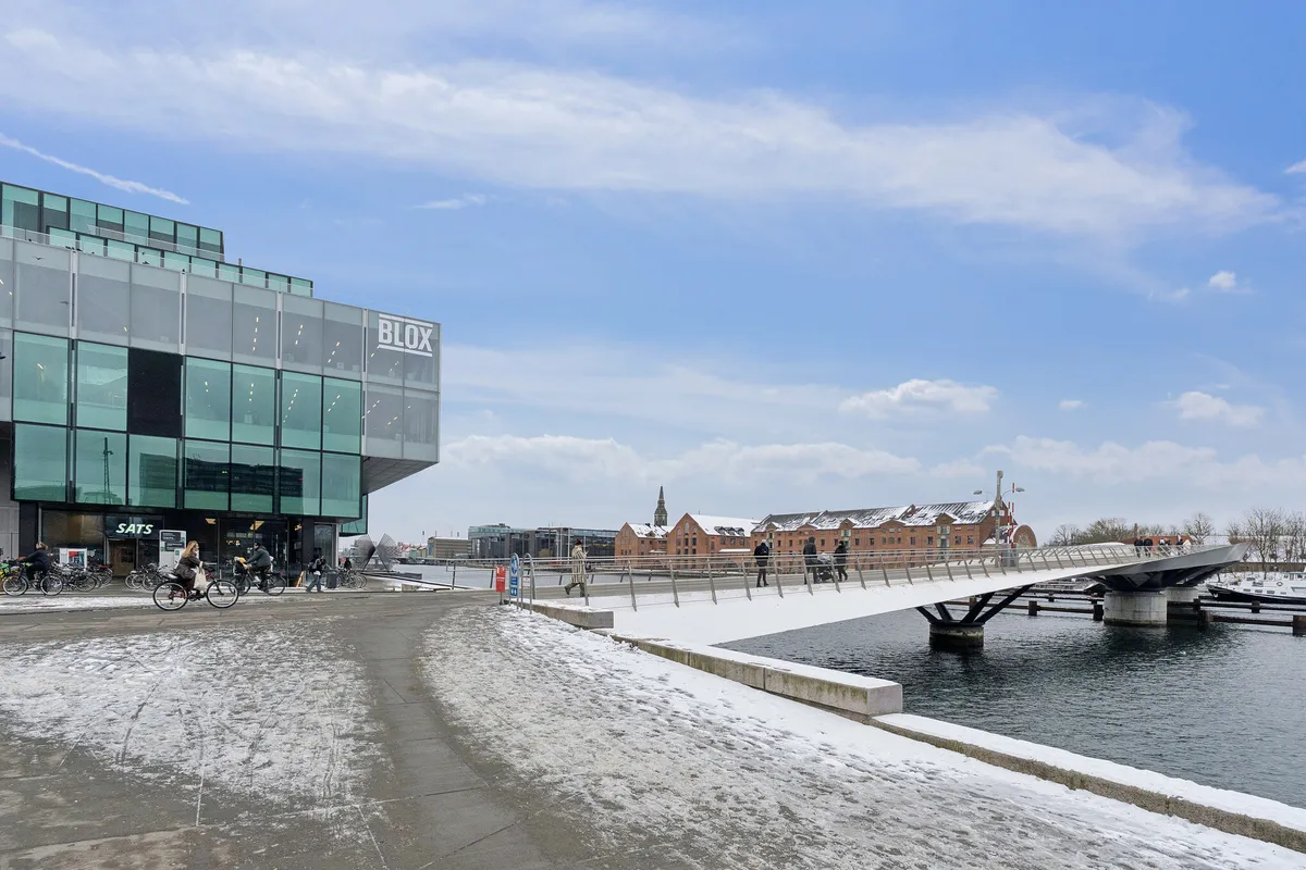 Mennesker cykler og går på en bro ved et bygningskompleks med navnet Blox i København. Slette veje og vandløb er dækket af sne. Bygninger og kanaler er i baggrunden